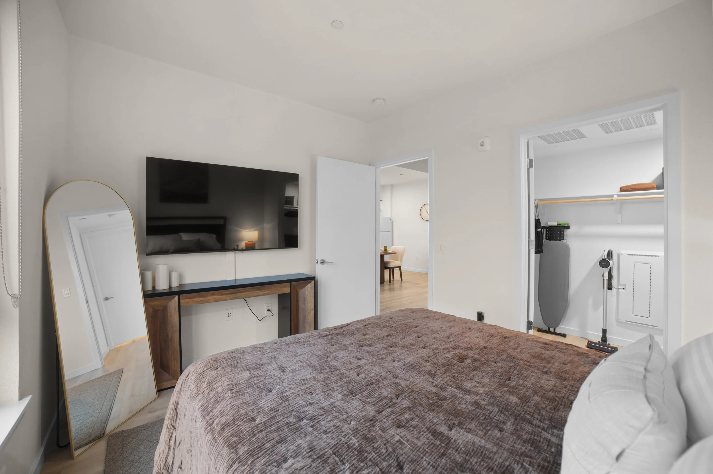 Interior view of a bedroom with a bed, flat-screen TV, a mirror leaning against the wall, and a doorway leading to a dining area. A closet with an iron and vacuum cleaner is visible.