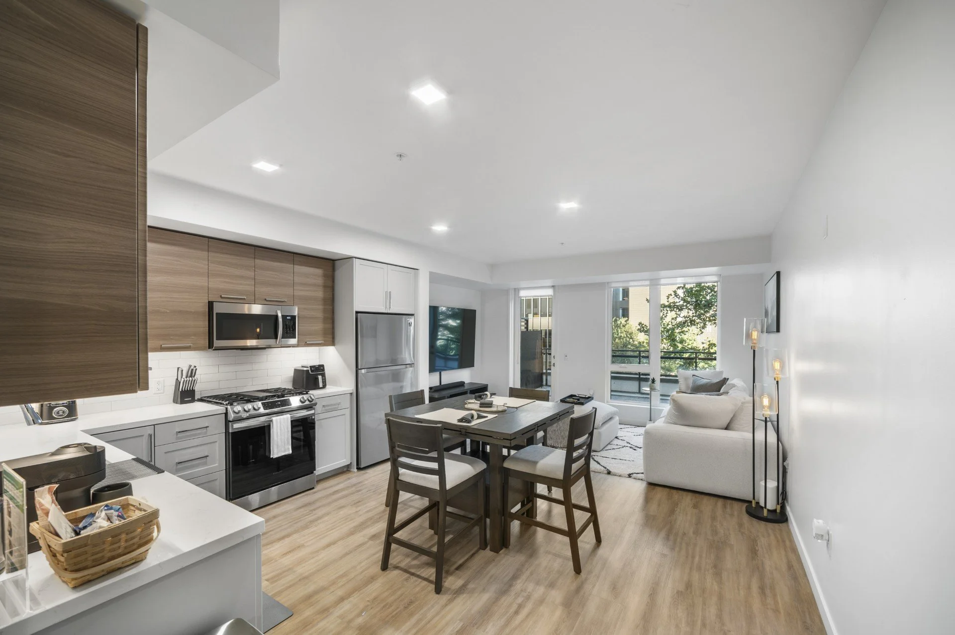 Interior view of a modern living room and kitchen with wooden floors, white walls, and large windows with trees outside. The kitchen has stainless steel appliances, including a refrigerator, microwave, and oven, with white and wood cabinets. There is