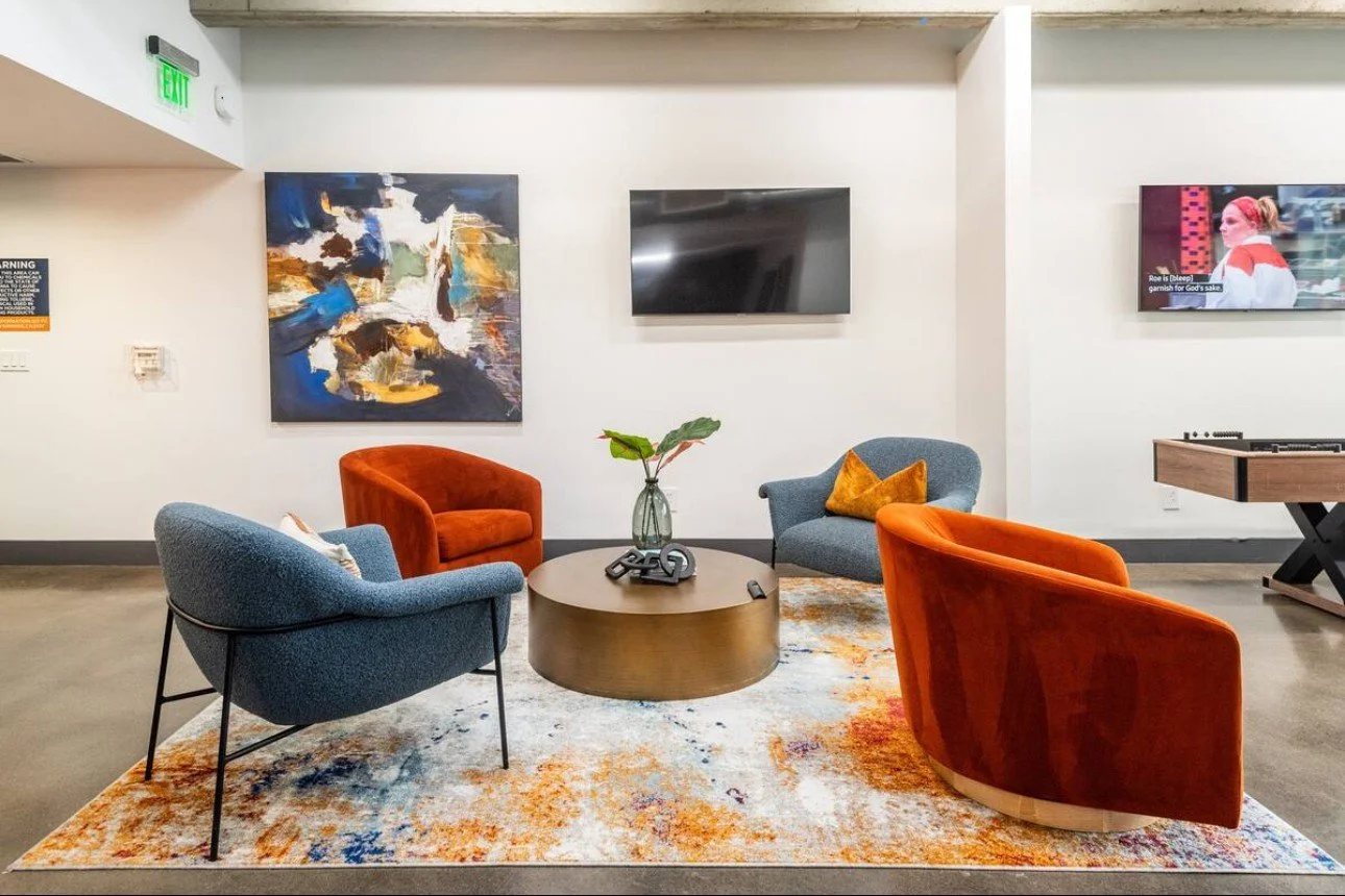 Modern lounge area with four colorful chairs - two orange and two blue - surrounding a round wooden table with a decorative black sculpture and a small plant. The area is on a patterned rug with abstract art on the wall, a large TV, a mounted flat-sc