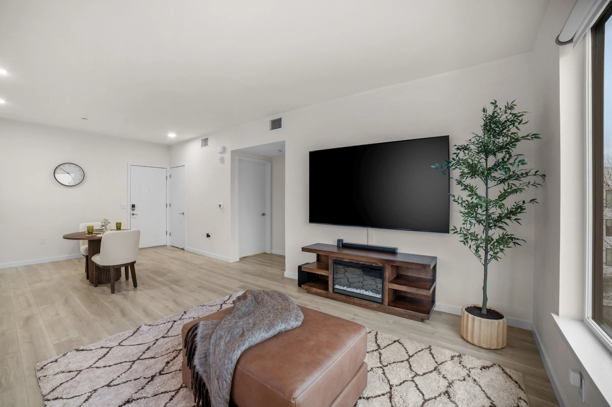 Living room with a mounted flat-screen TV, wooden TV stand, a potted plant, and a brown leather ottoman on a patterned rug. A dining area with a small round table and four white chairs is in the background, with a wall clock visible.