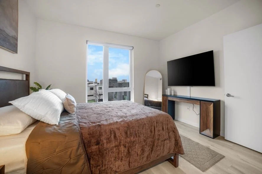 Modern bedroom with a large window, bed with brown bedding, flat-screen TV on the wall, and a mirror near the door.