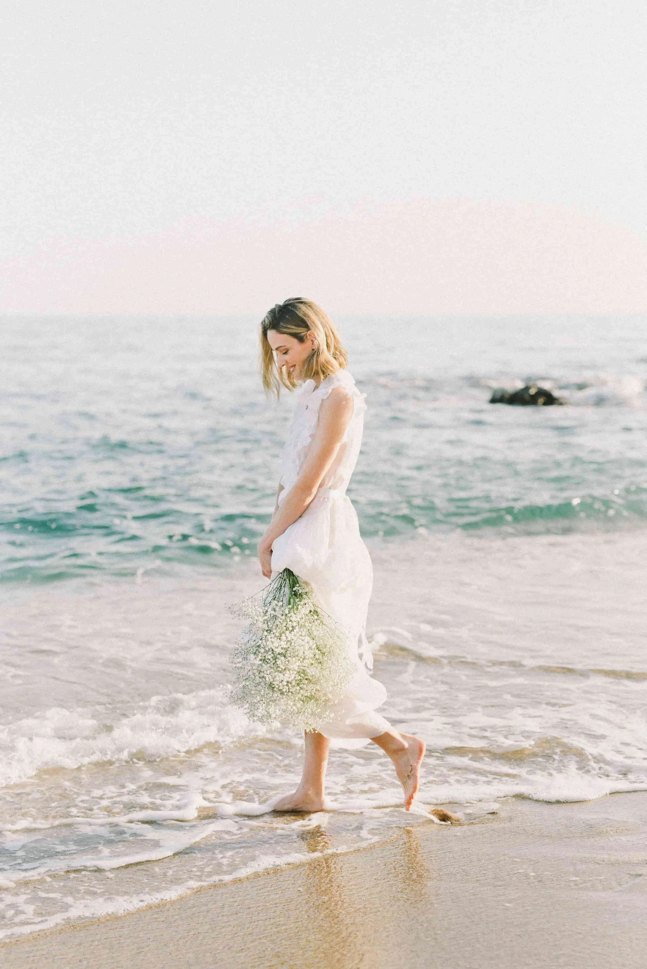Woman in a white dress walking in the ocean, holding a bouquet of flowers, with waves and rocks in the background.