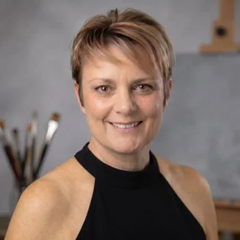 A woman with short hair smiling in a black sleeveless top, standing in front of a gray wall with paintbrushes in the background.