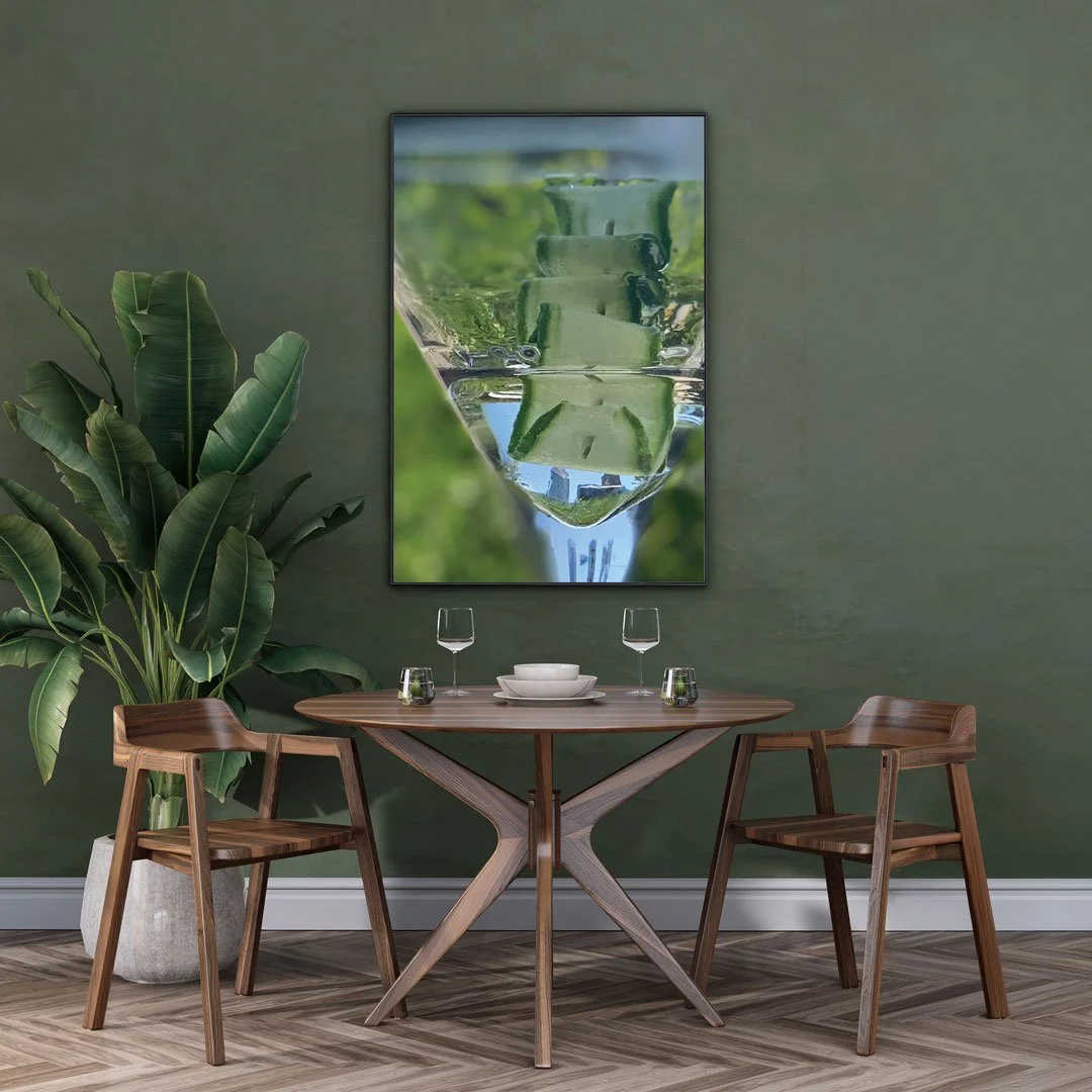 A restaurant setting featuring a round wooden table set with wine glasses, bowls, and cups, surrounded by two wooden chairs, with a large potted plant on the left and a green wall with a reflection artwork on the background.