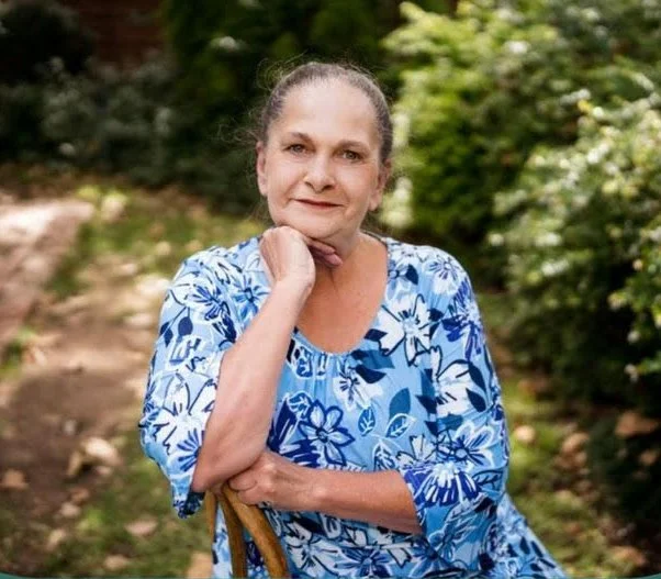 A woman with gray hair in a bun, wearing a blue and white floral dress, sitting outdoors with greenery in the background.