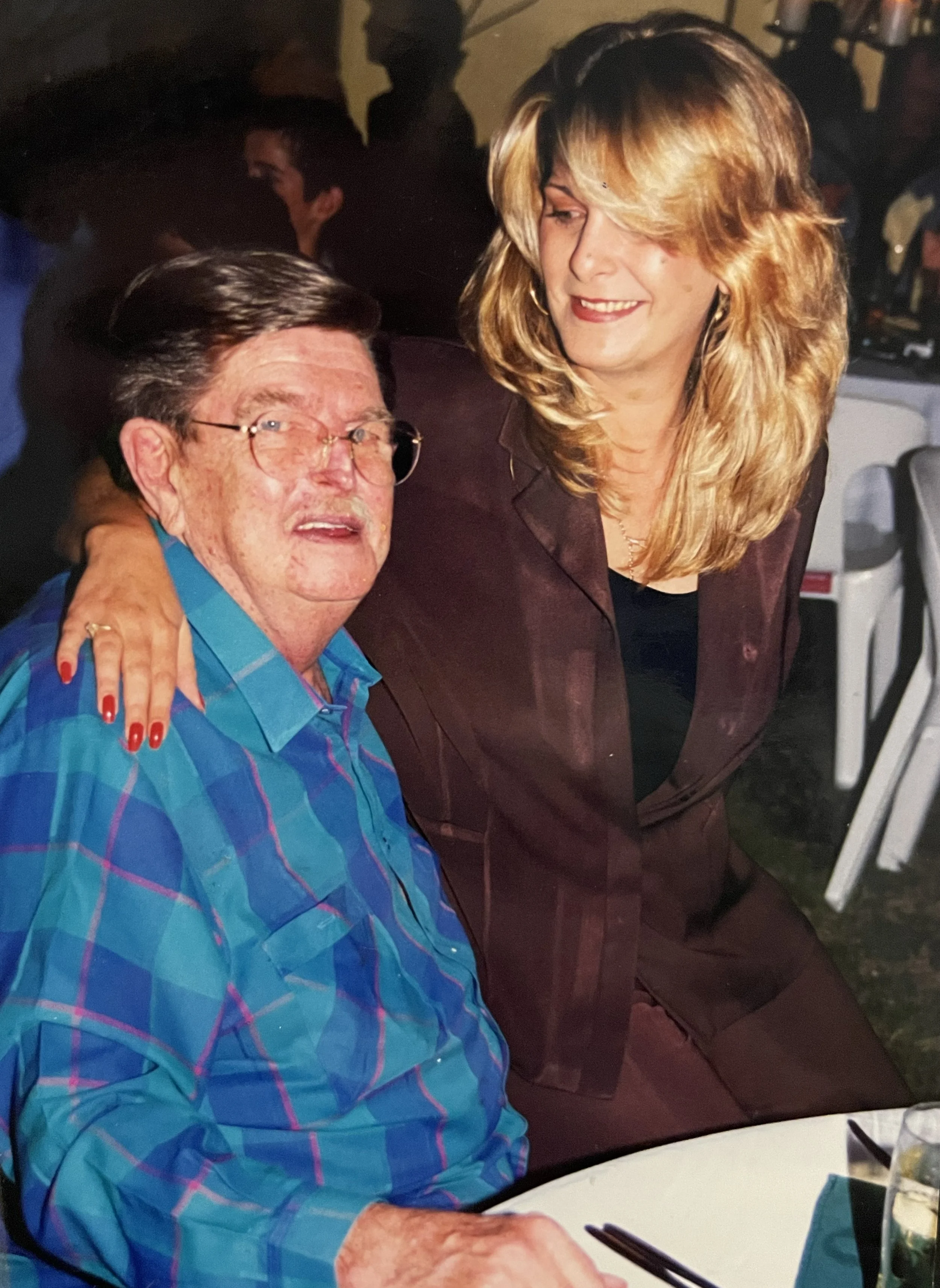A woman with blonde hair and a man wearing glasses and a blue plaid shirt pose together at a social event, smiling with the woman's arm around the man's shoulders.