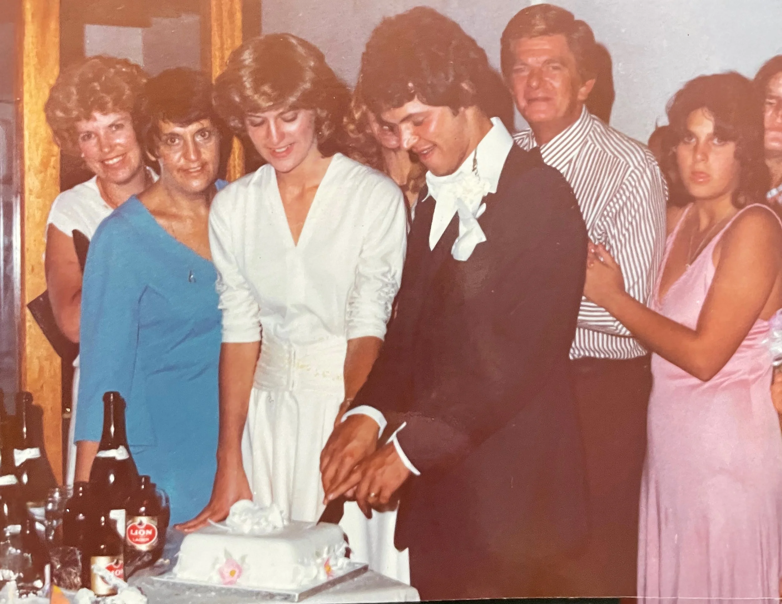 A group of people celebrating around a cake, with two bottles of beer and some glasses on the table, in a festive indoor setting.