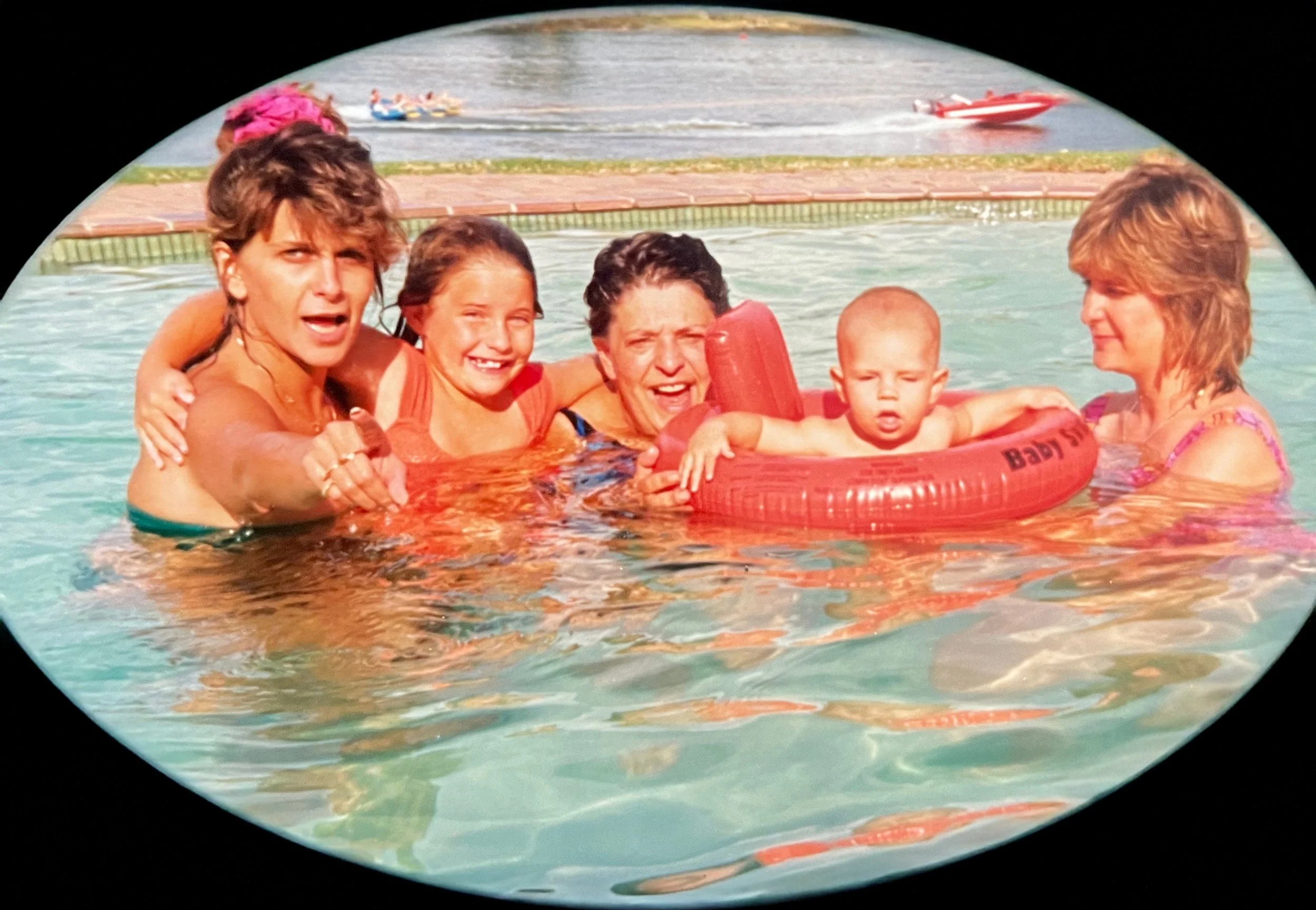 Five women and a baby in a swimming pool, with children’s toys and a dock in the background.