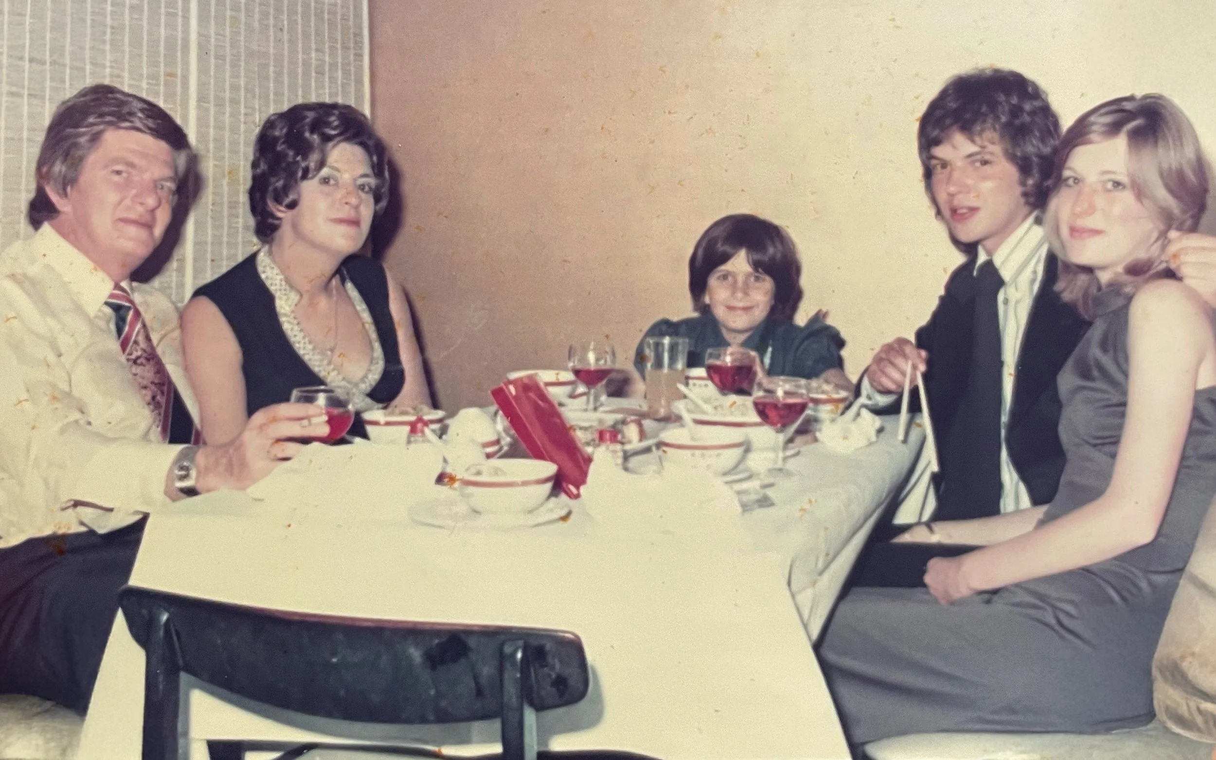 Six people sitting at a dining table with bowls, glasses of wine, and a red box. They are dressed formally and smiling at the camera.