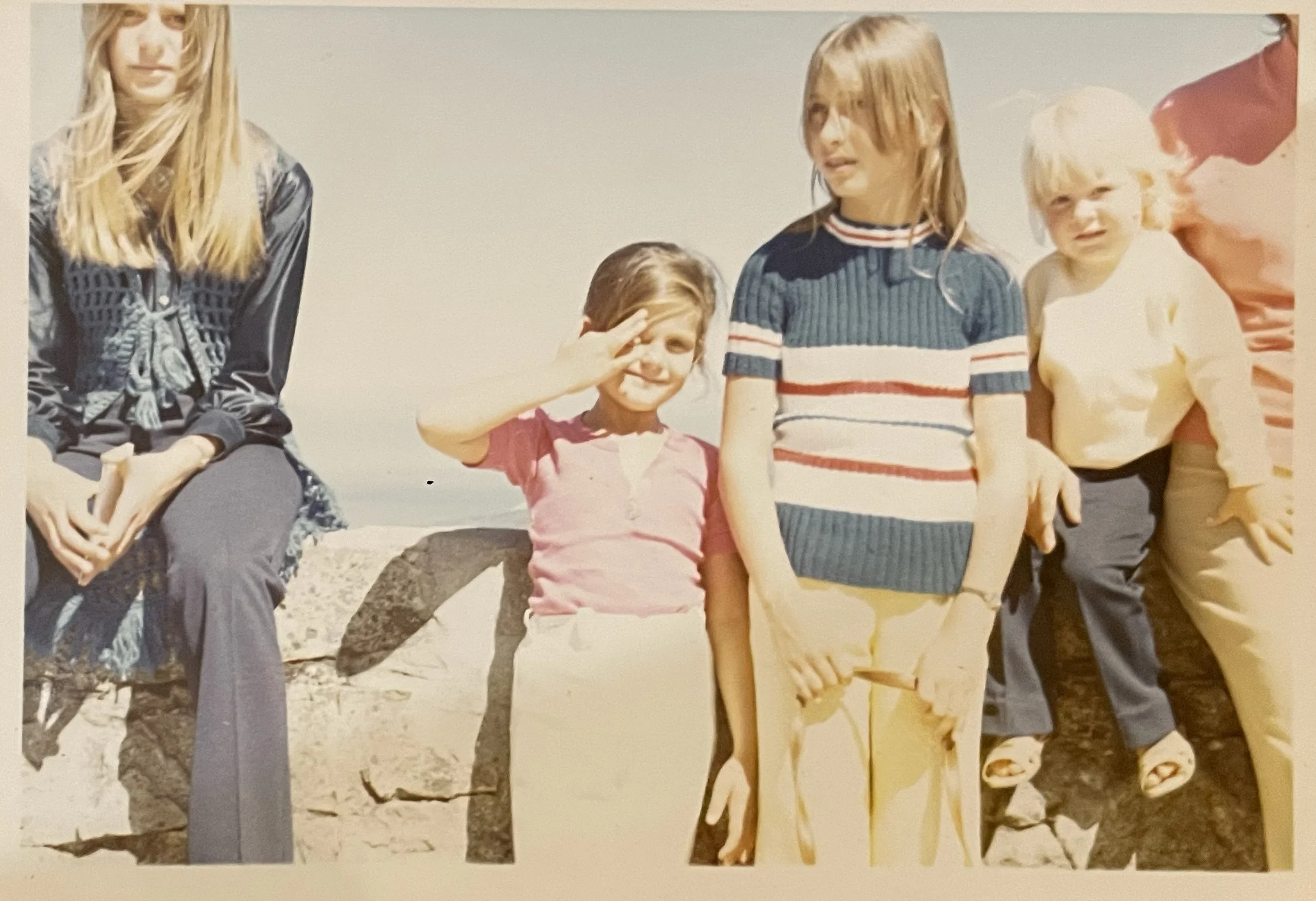 A group of four children sitting outdoors on rocks, with two girls standing and two boys sitting behind, during daytime with a clear sky.