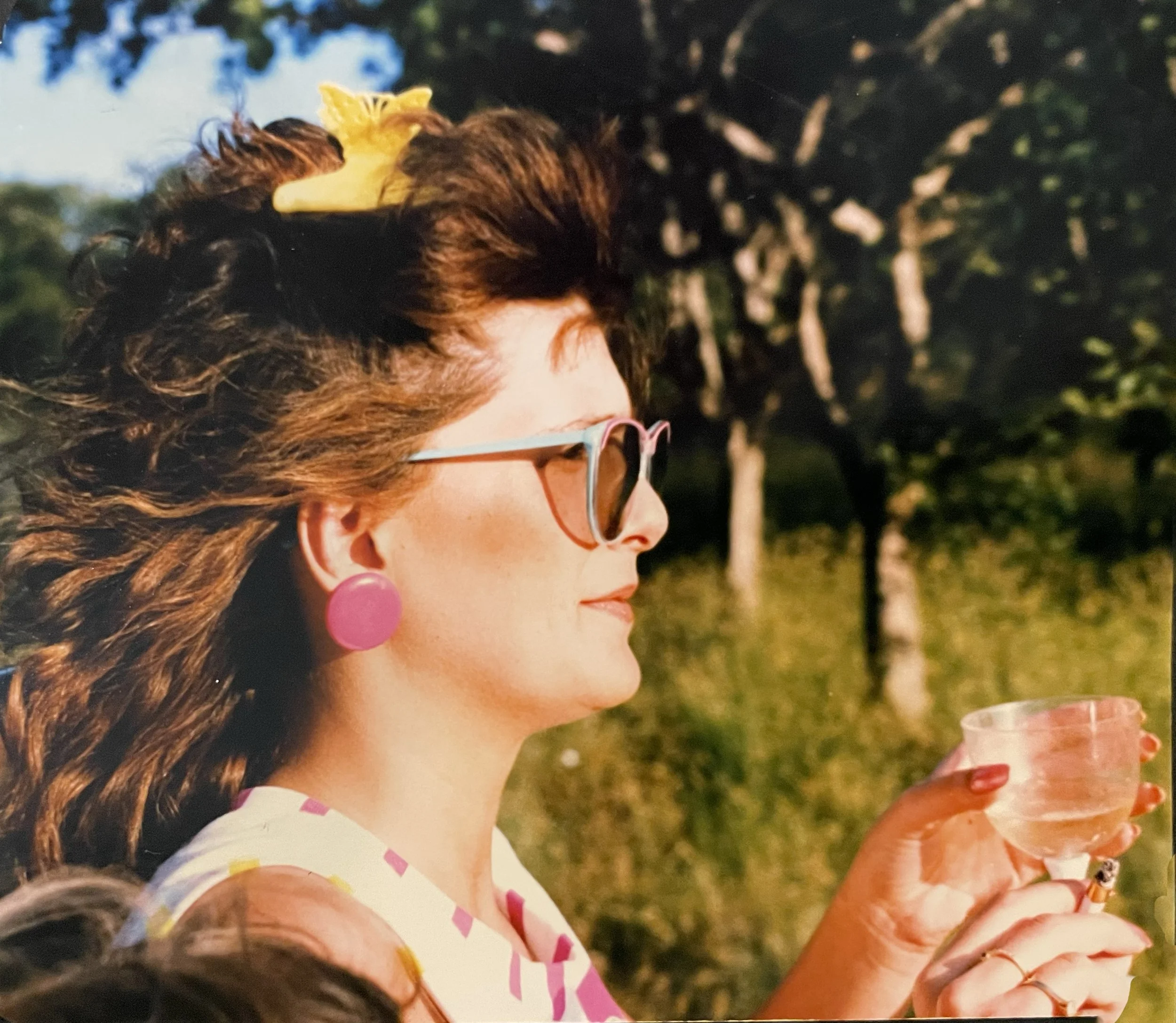 A woman with curly brown hair, wearing pink earrings, sunglasses, and a white shirt with pink accents. She is holding a glass cup outdoors during daytime, with trees in the background.