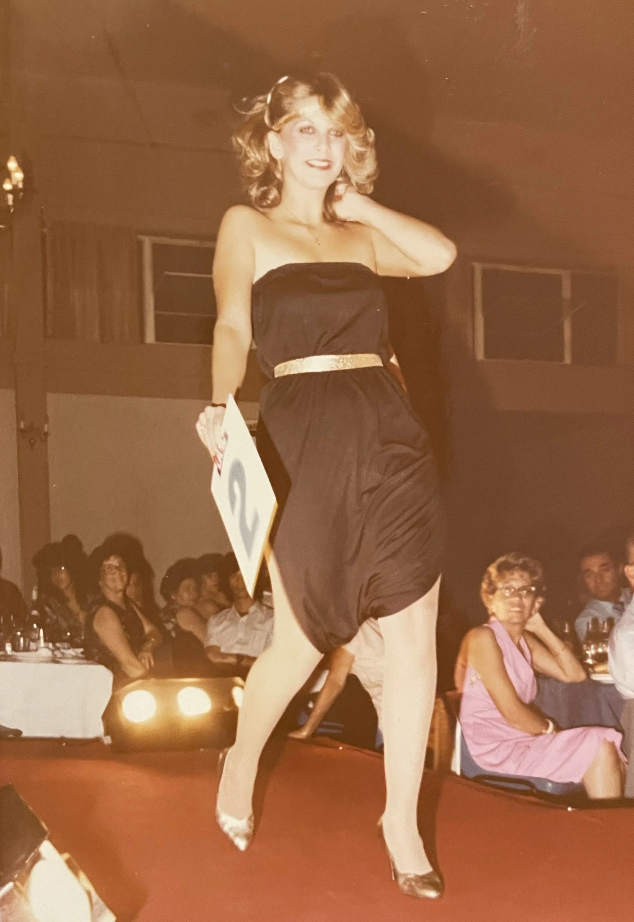 A woman in a black strapless dress with a gold belt is dancing on a stage, holding a number two card, with an audience watching in the background.