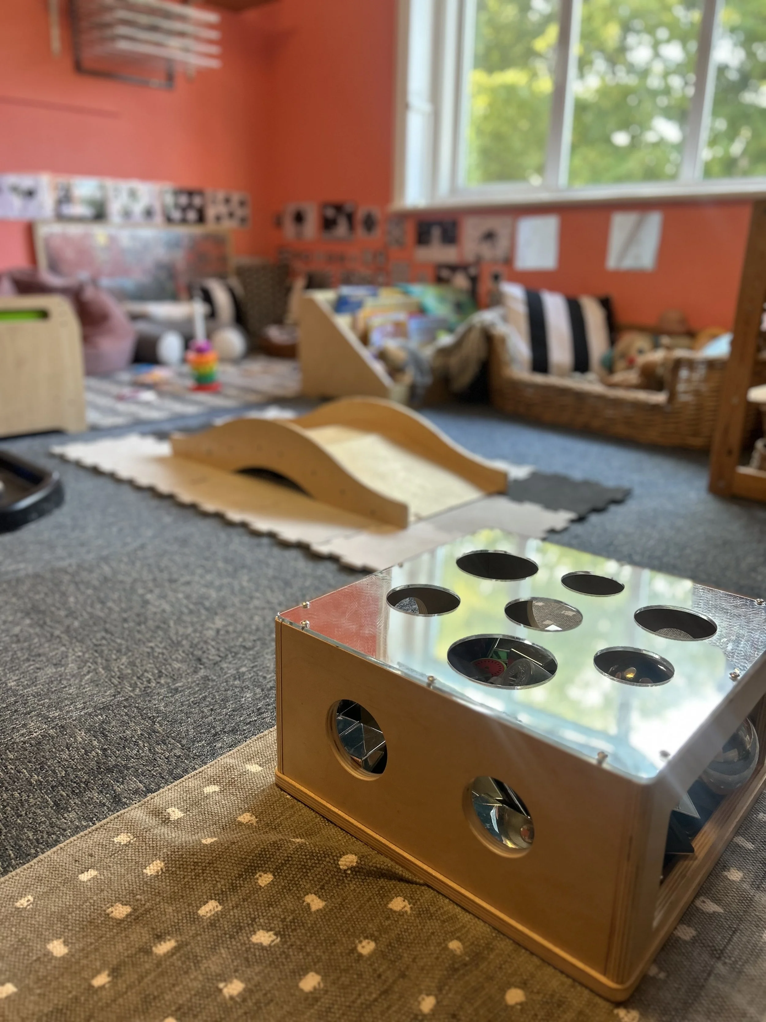 A wooden box with a mirrored top and round holes, placed on a carpeted floor in a children's playroom. The room contains a small slide, toys, pet beds, and photographs on the walls, with a large window letting in natural light.