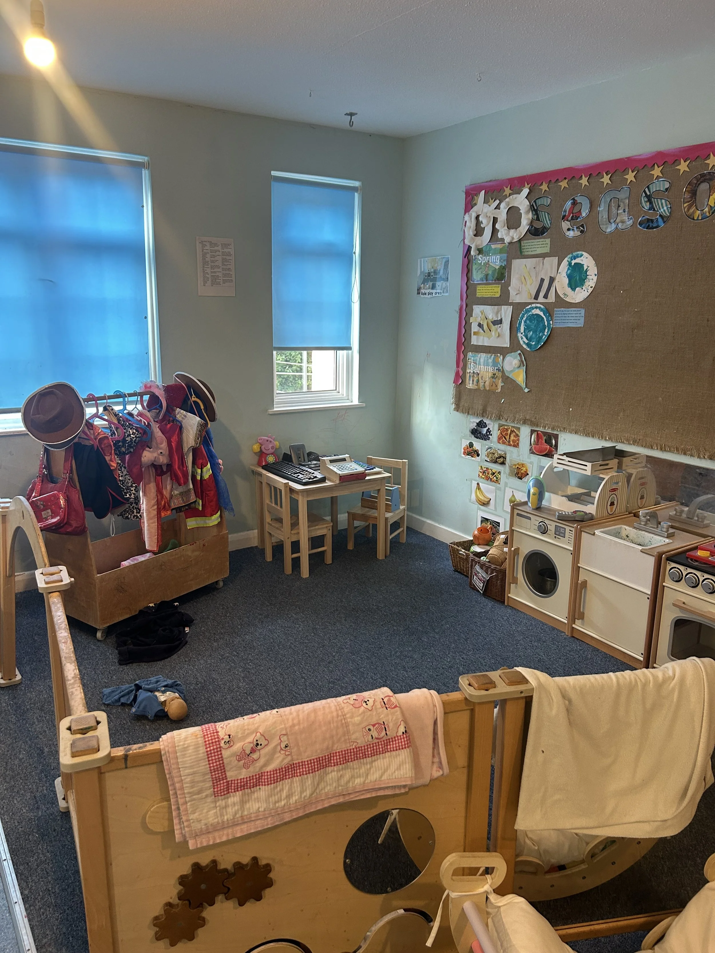 Empty preschool classroom with coat rack, play kitchen, educational posters, and small tables and chairs.