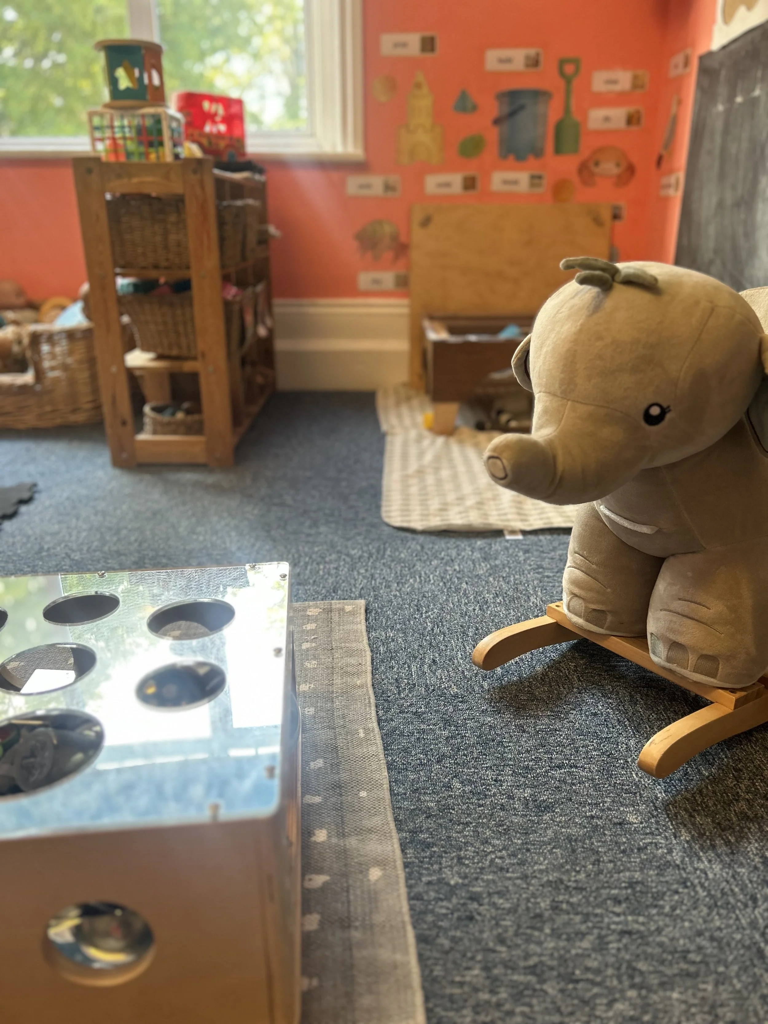 A plush toy pig on a wooden rocking stand in a children's playroom with colorful walls and shelves filled with toys.