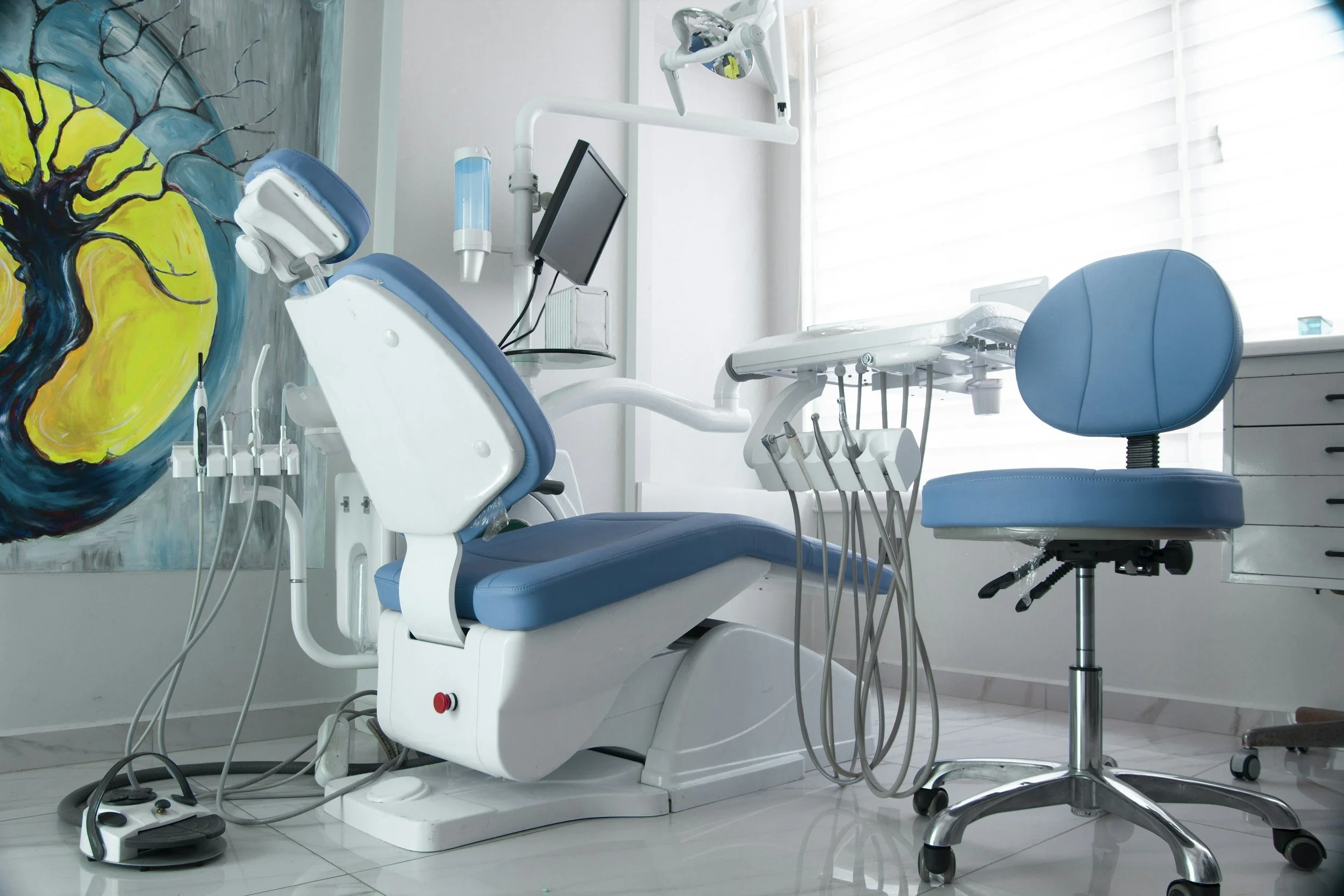 An empty dental examination room showing a blue dental chair, a blue rolling stool, dental tools, a monitor, and a colorful abstract wall painting.