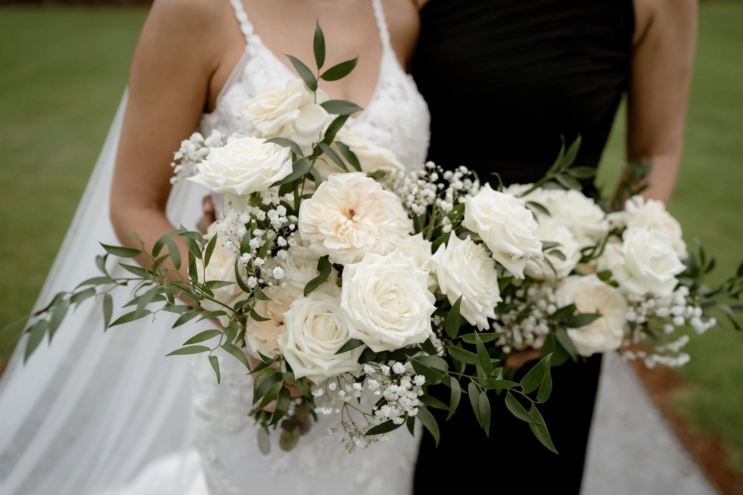 Two women holding large bouquets of white roses, baby's breath, and greenery during a wedding.