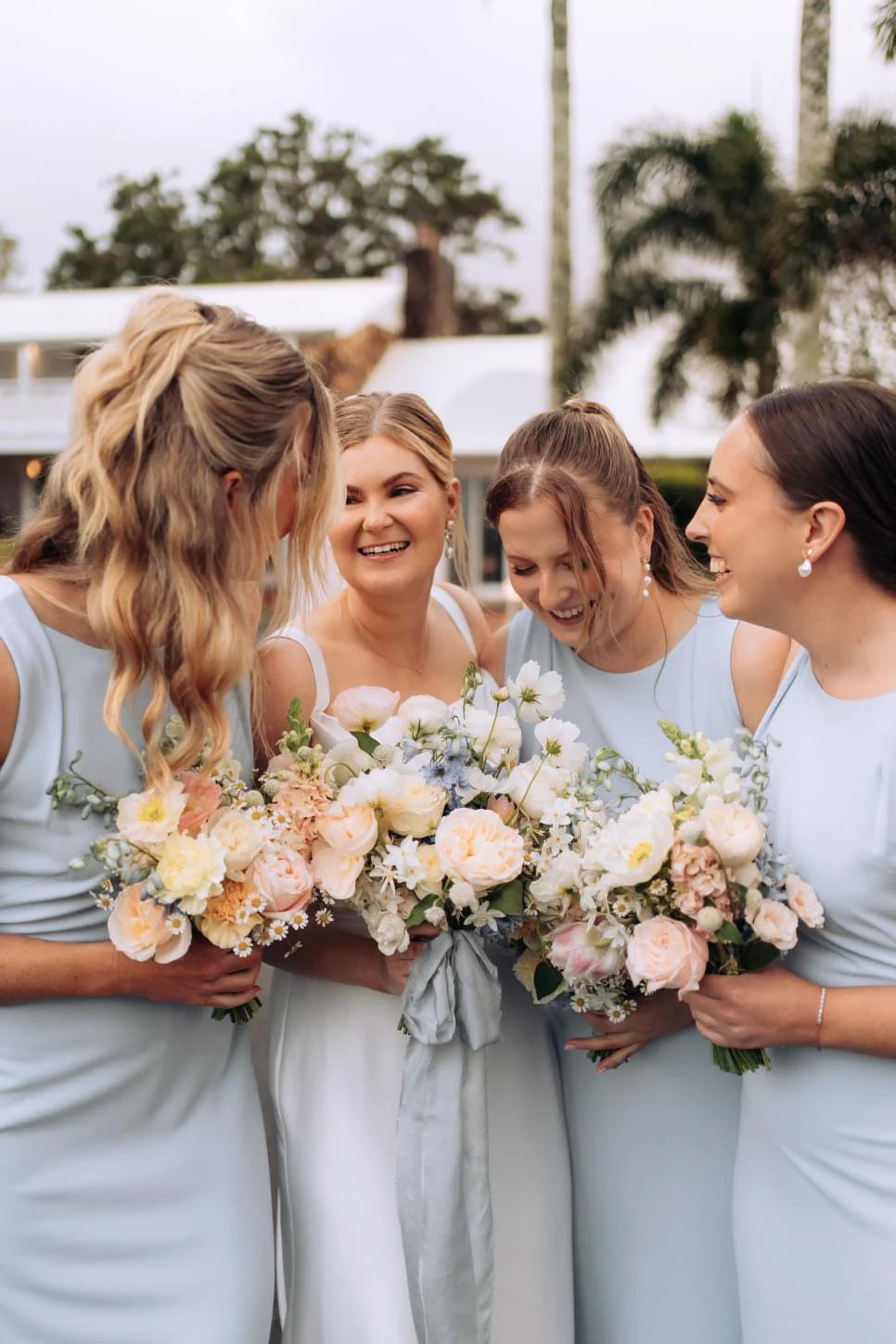 Four women in light blue dresses holding bouquets, smiling and laughing together outdoors.