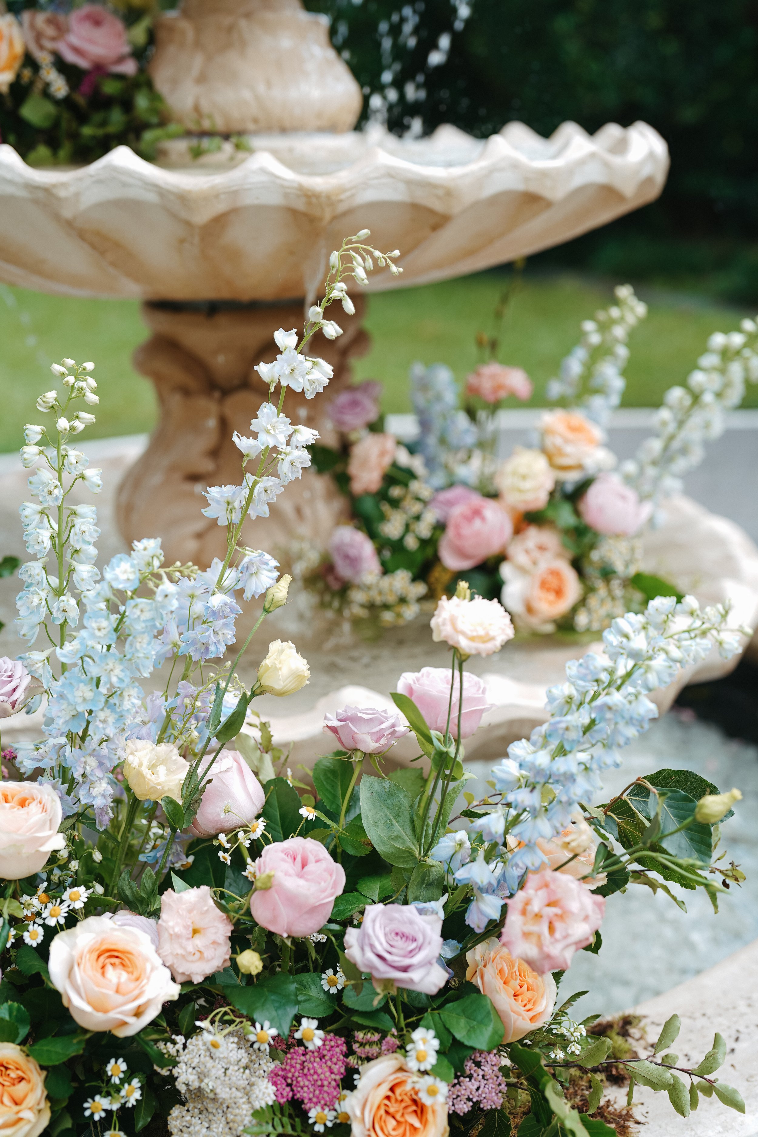 A floral arrangement with pink and white roses, blue delphiniums, and small white daisies in front of a two-tiered cream-colored stone fountain.