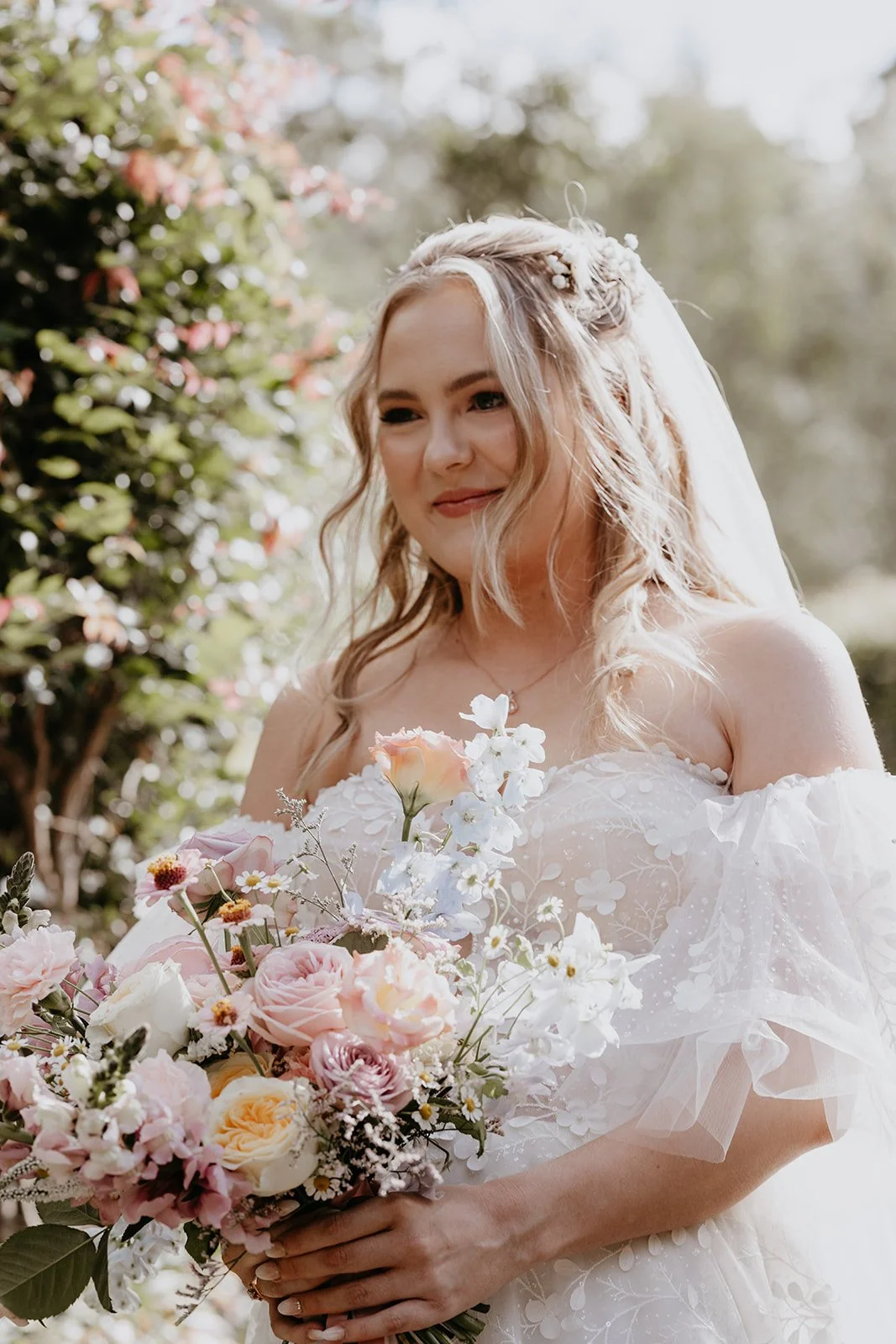 A bride in a white wedding dress holding a bouquet of pink, white, and peach roses with greenery outdoors.