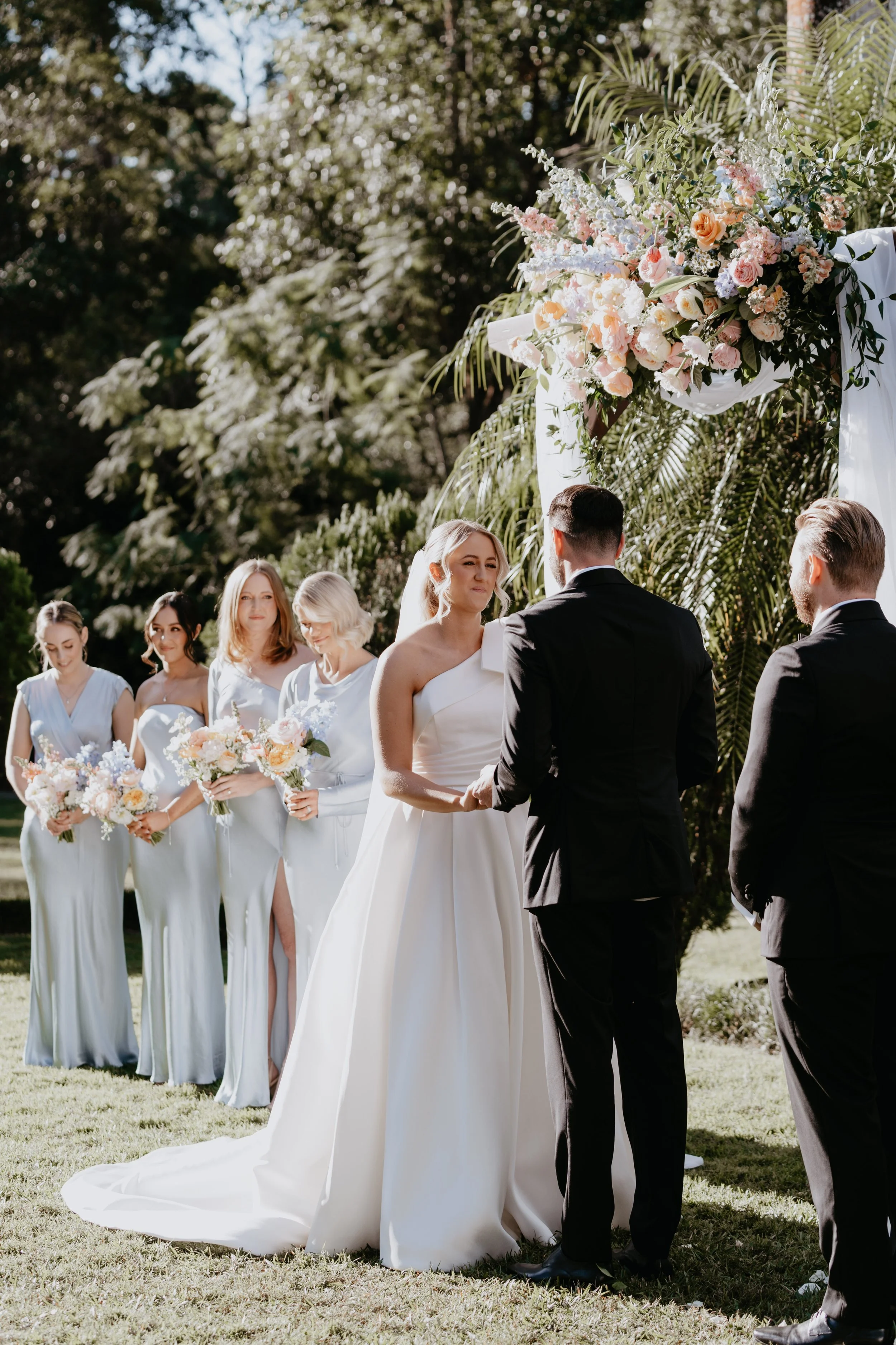 Bride and groom exchanging vows outdoors with bridesmaids and groomsmen in the background and a floral arch.