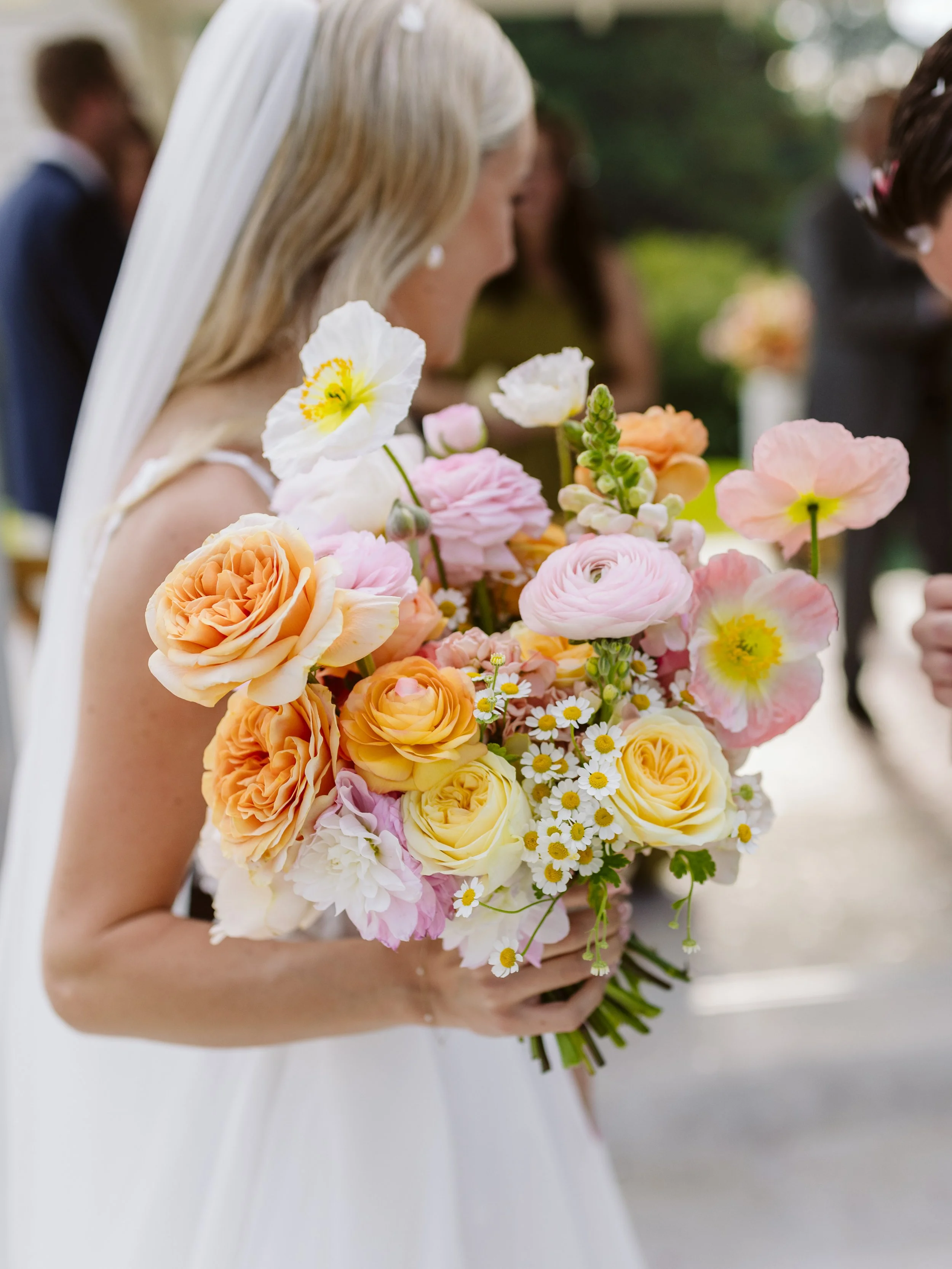 A bride holding a large bouquet of pink, peach, yellow, and white flowers at a wedding.