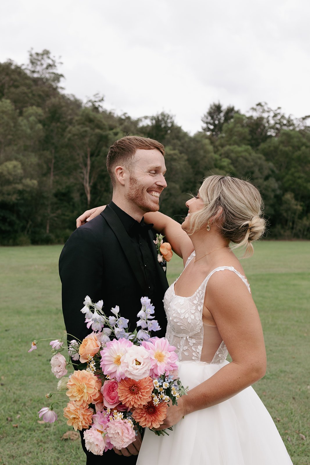 A newlywed couple smiling and hugging outdoors, the bride holding a bouquet of pink, peach, and white flowers, with green trees and grass in the background.