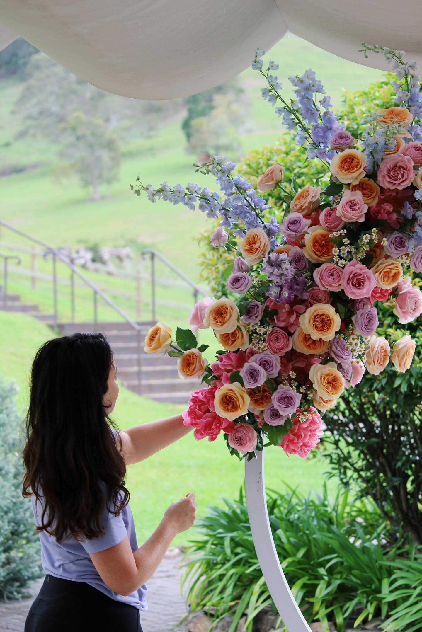 A woman arranging a large colorful floral display outdoors with a green landscape and stairs in the background.