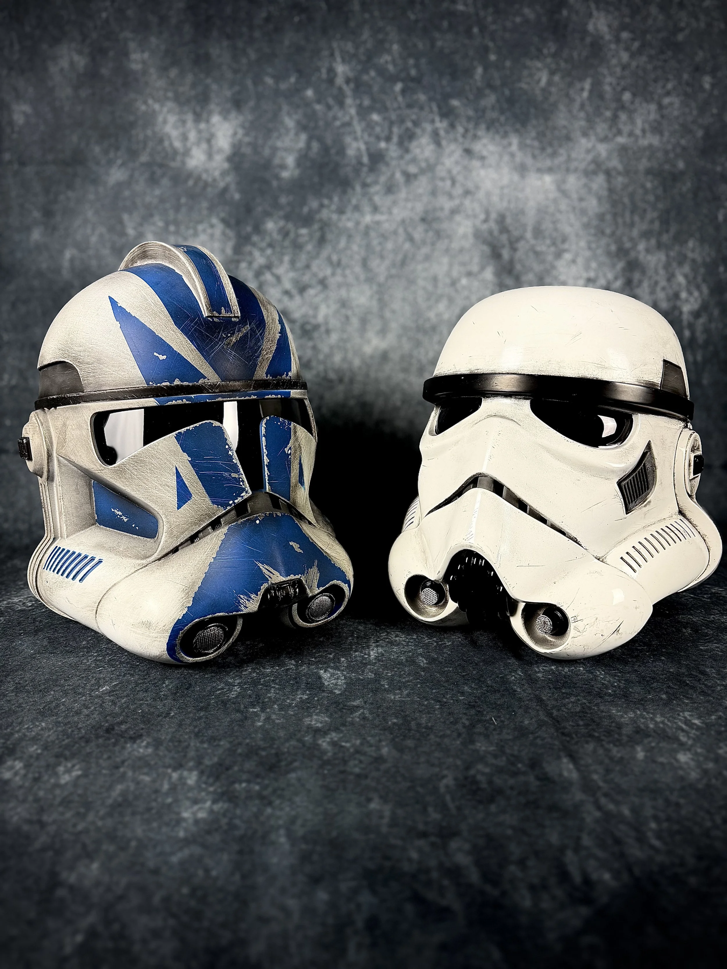Two Star Wars stormtrooper helmets, one with blue and white paint and the other plain white, placed on a dark textured surface with a dark background.