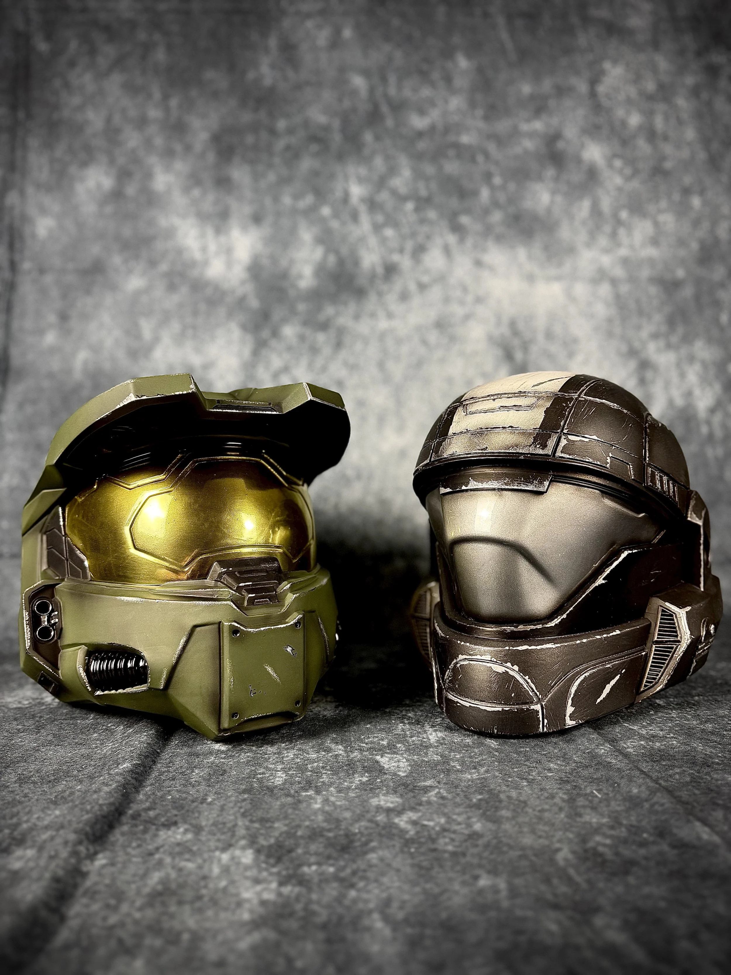 Two futuristic helmets, one with a green and gold design and the other with a black and silver design, placed on a gray textured surface with a gray background.