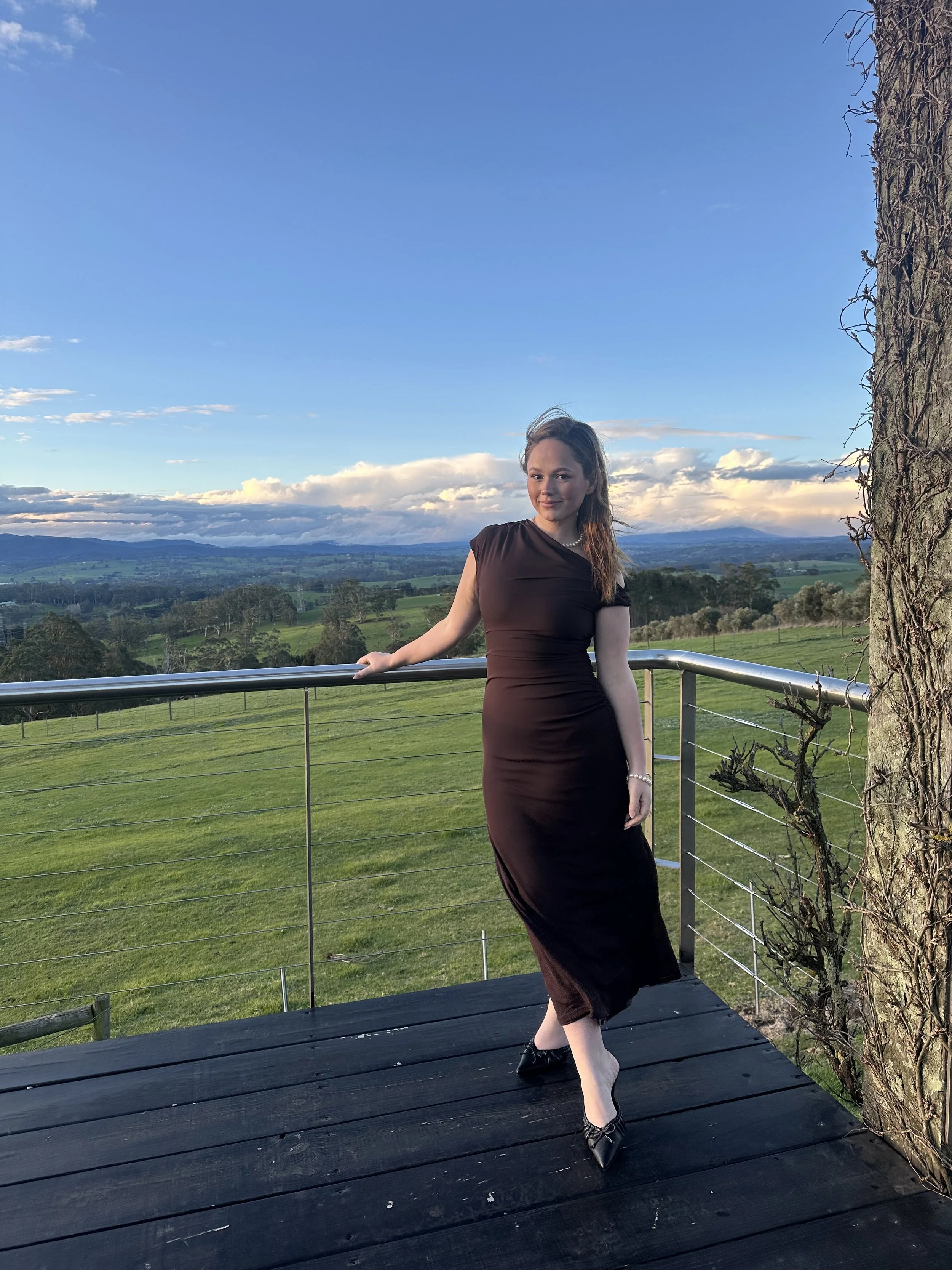 A woman in a black dress standing on a wooden deck by a metal railing, overlooking a green landscape with fields, trees, and mountains in the distance during sunset.