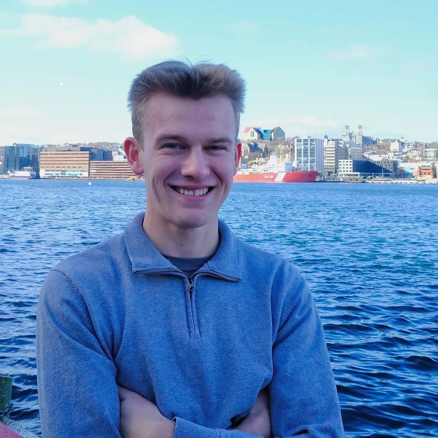 Young man smiling with arms crossed in front of a harbor with buildings and a ship in the background.