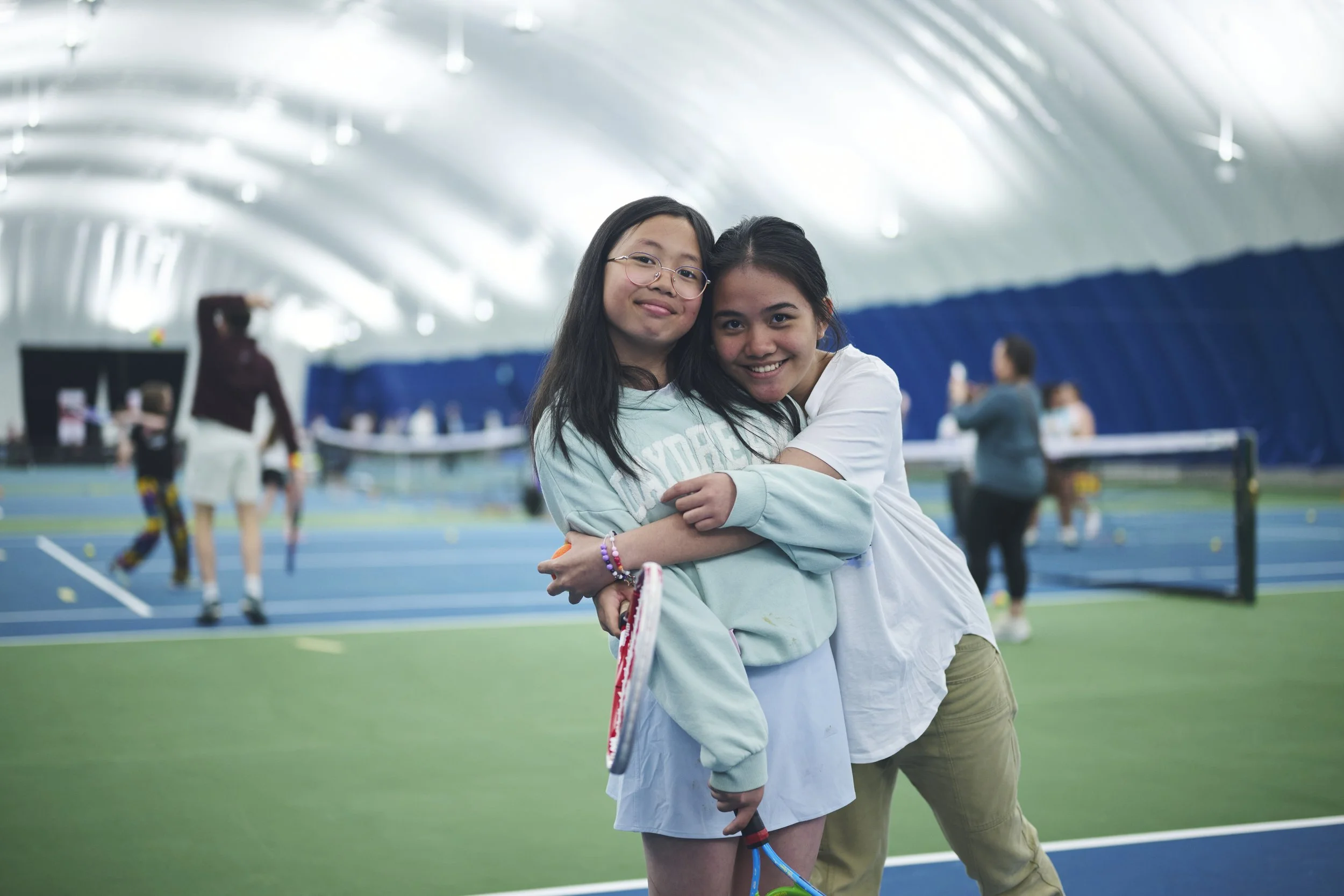 Two young girls hugging inside an indoor tennis court, with people playing tennis in the background.