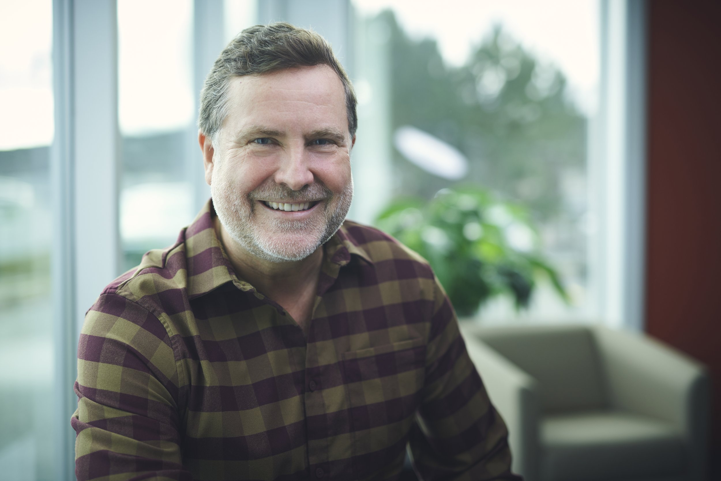 A middle-aged man with short brown hair and a beard, smiling, wearing a plaid shirt, sitting indoors near large windows with a blurred view of trees and a house outside, and a green plant in the background.