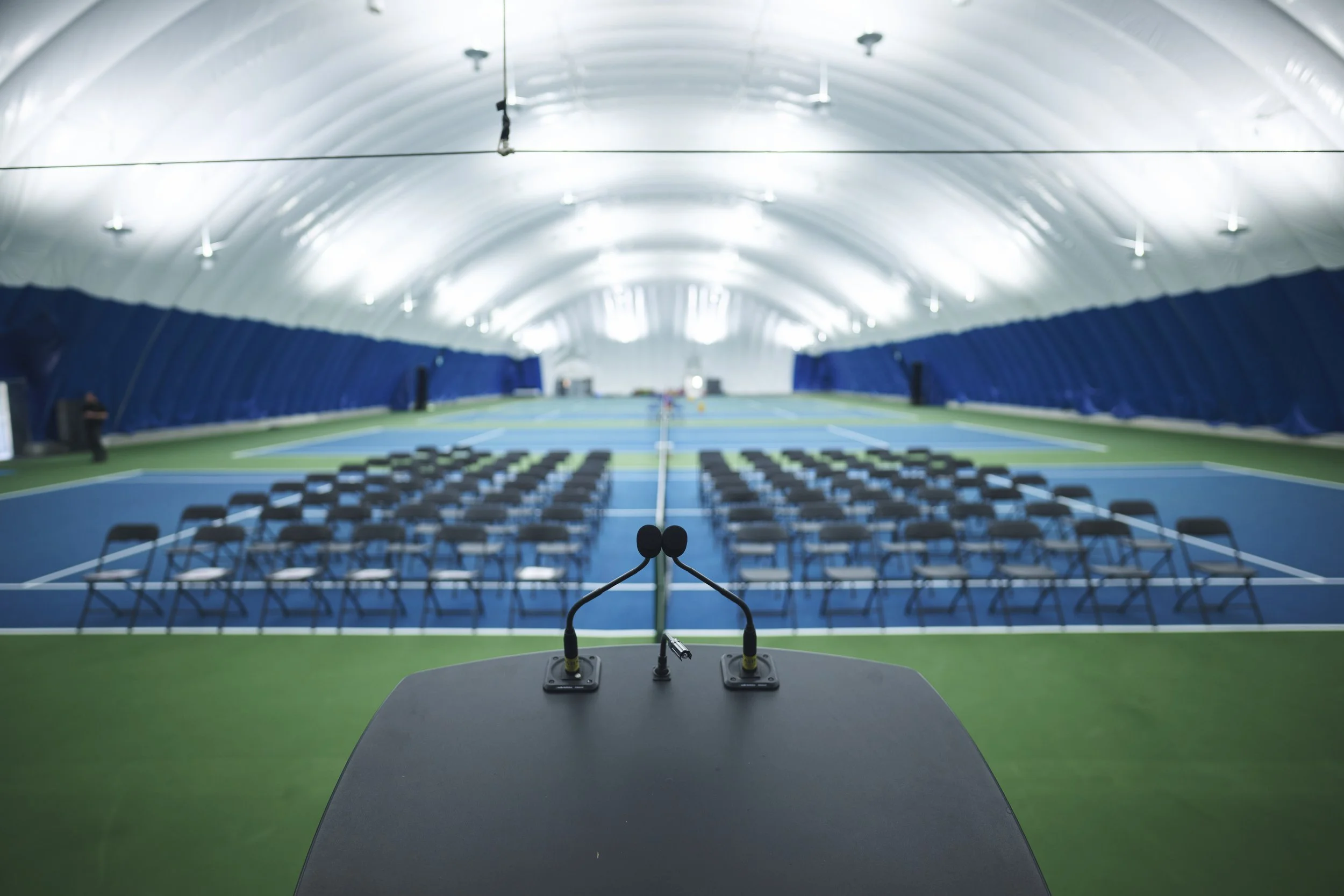 Empty indoor tennis court set up with chairs and a lectern, viewed from the podium.