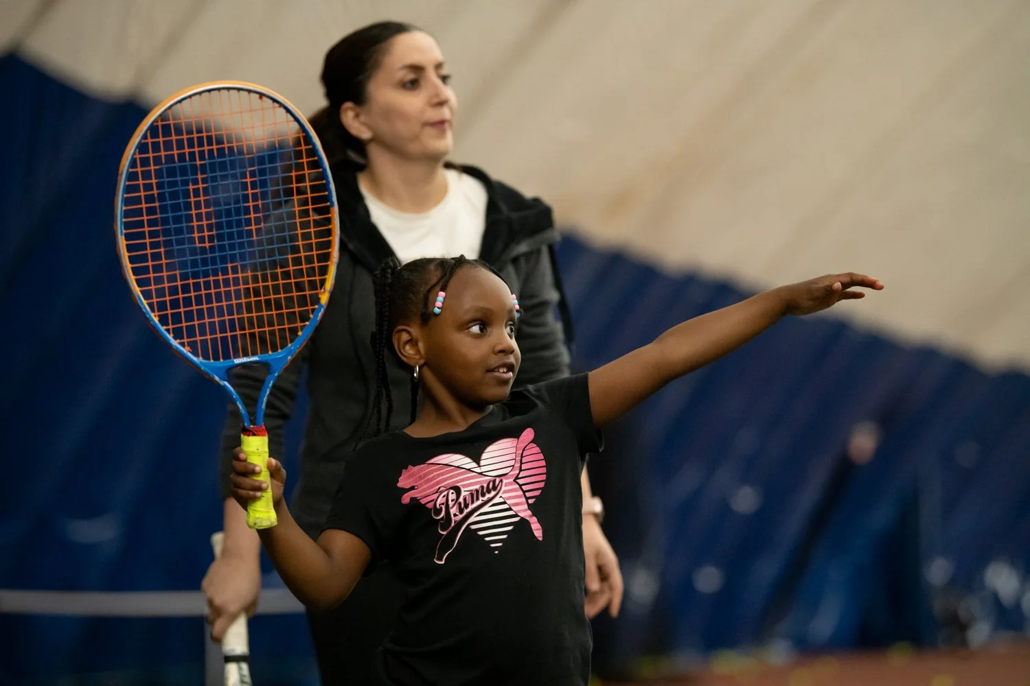 A young girl with braided hair and colorful beads holding a tennis racket, pointing forward with a woman standing behind her supervising indoors.