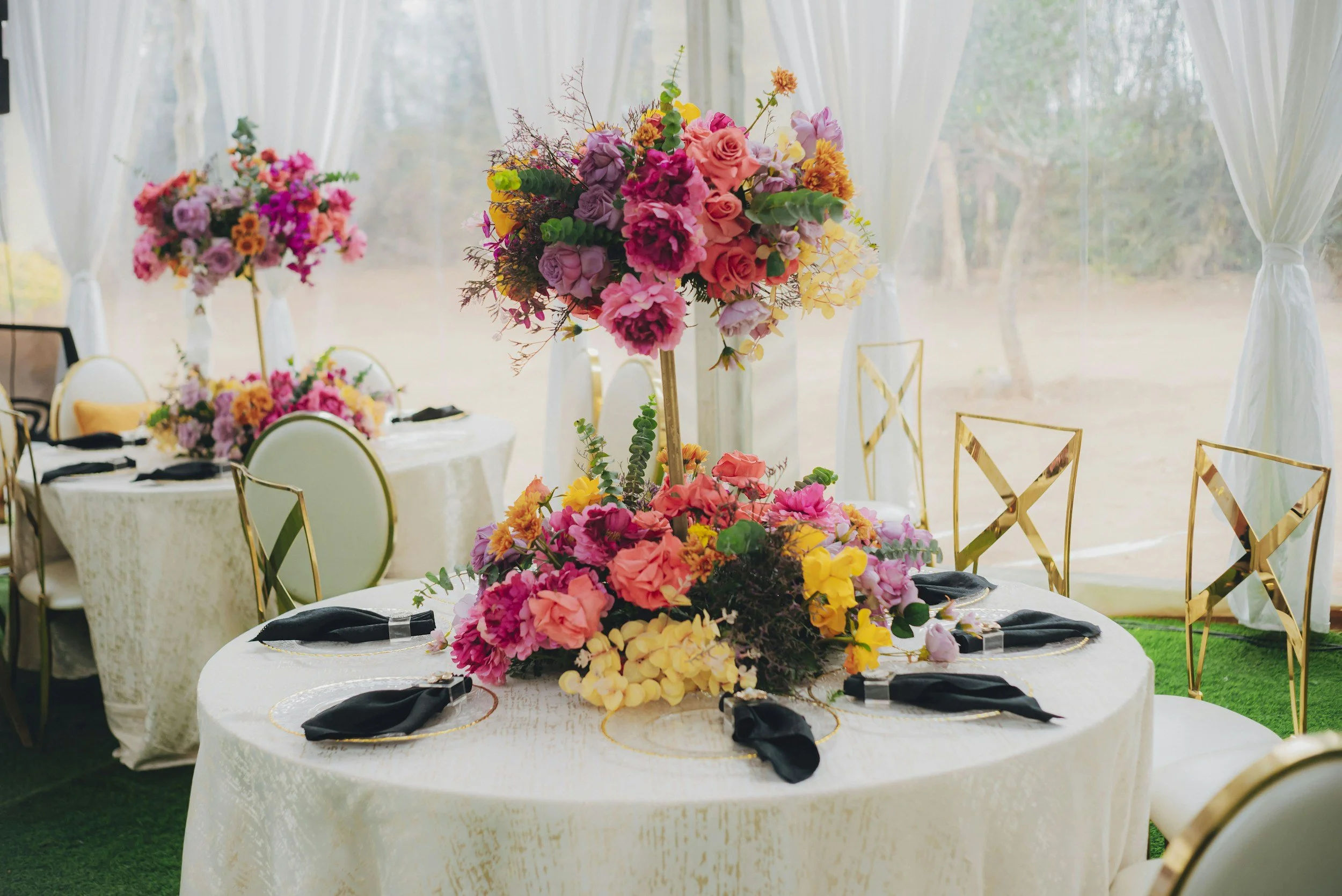 Elegant dining table decorated with a tall flower arrangement featuring pink and purple flowers, surrounded by gold and white chairs, with a backdrop of white curtains and outdoor scenery.