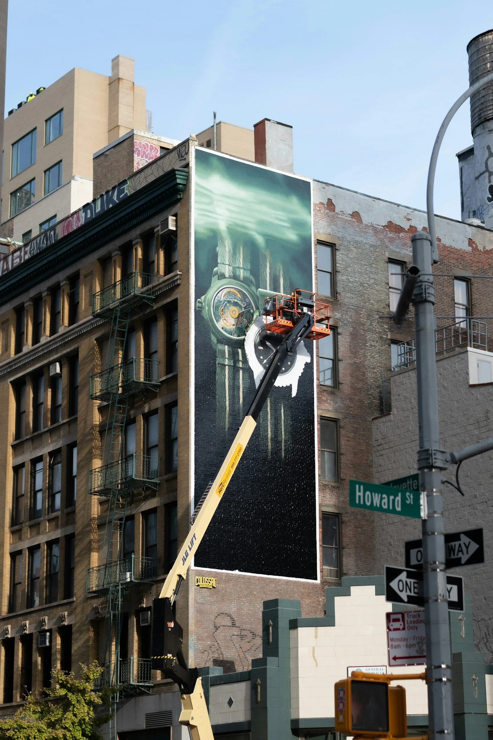 A large building with a mural of a clock and green liquid on the side, with a cherry picker lift in front, at the corner of Howard Street and Lafayette.