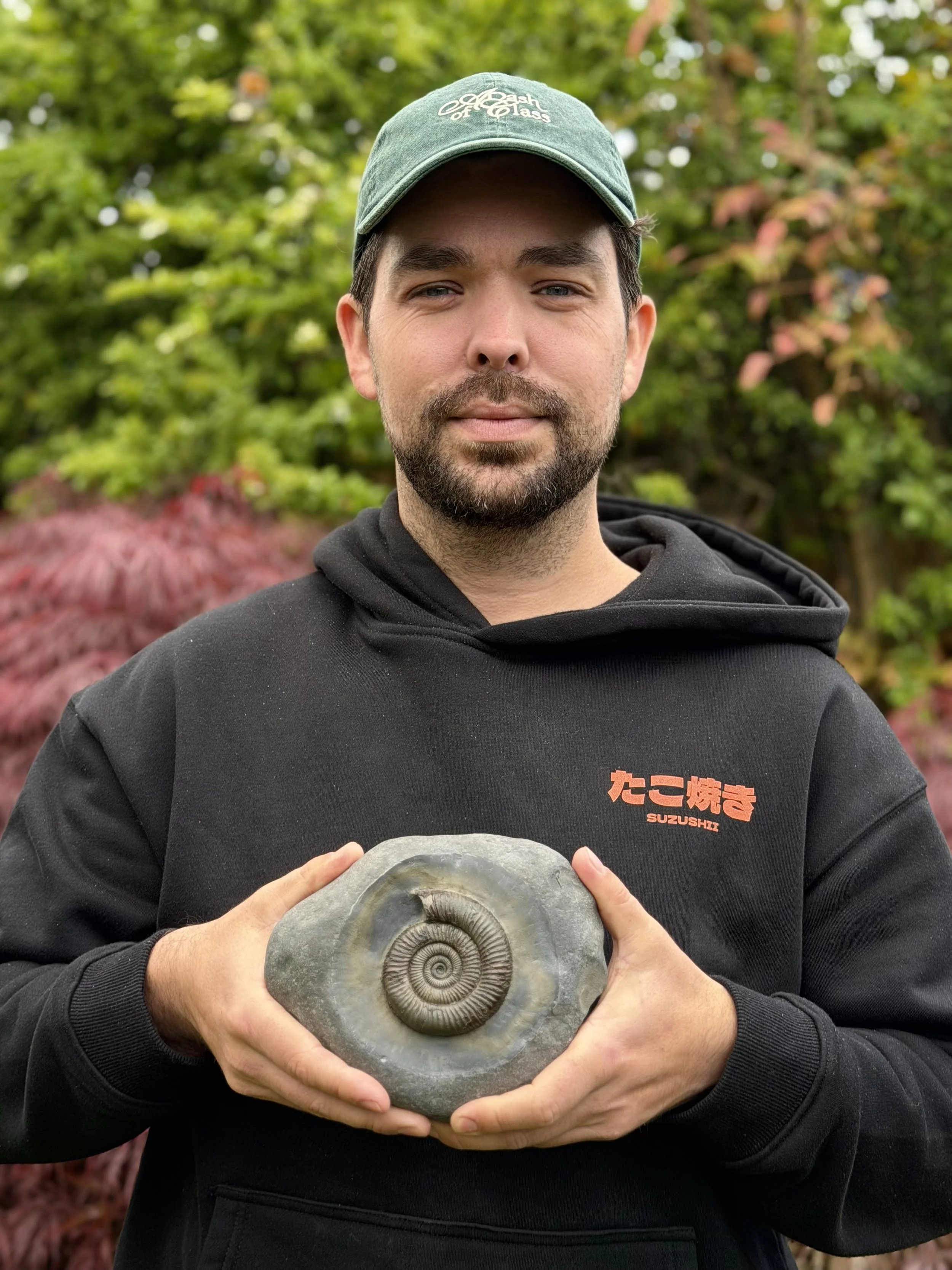 A man with a beard wearing a green baseball cap and a black hoodie holding a large ammonite fossil outdoors with trees and bushes in the background.
