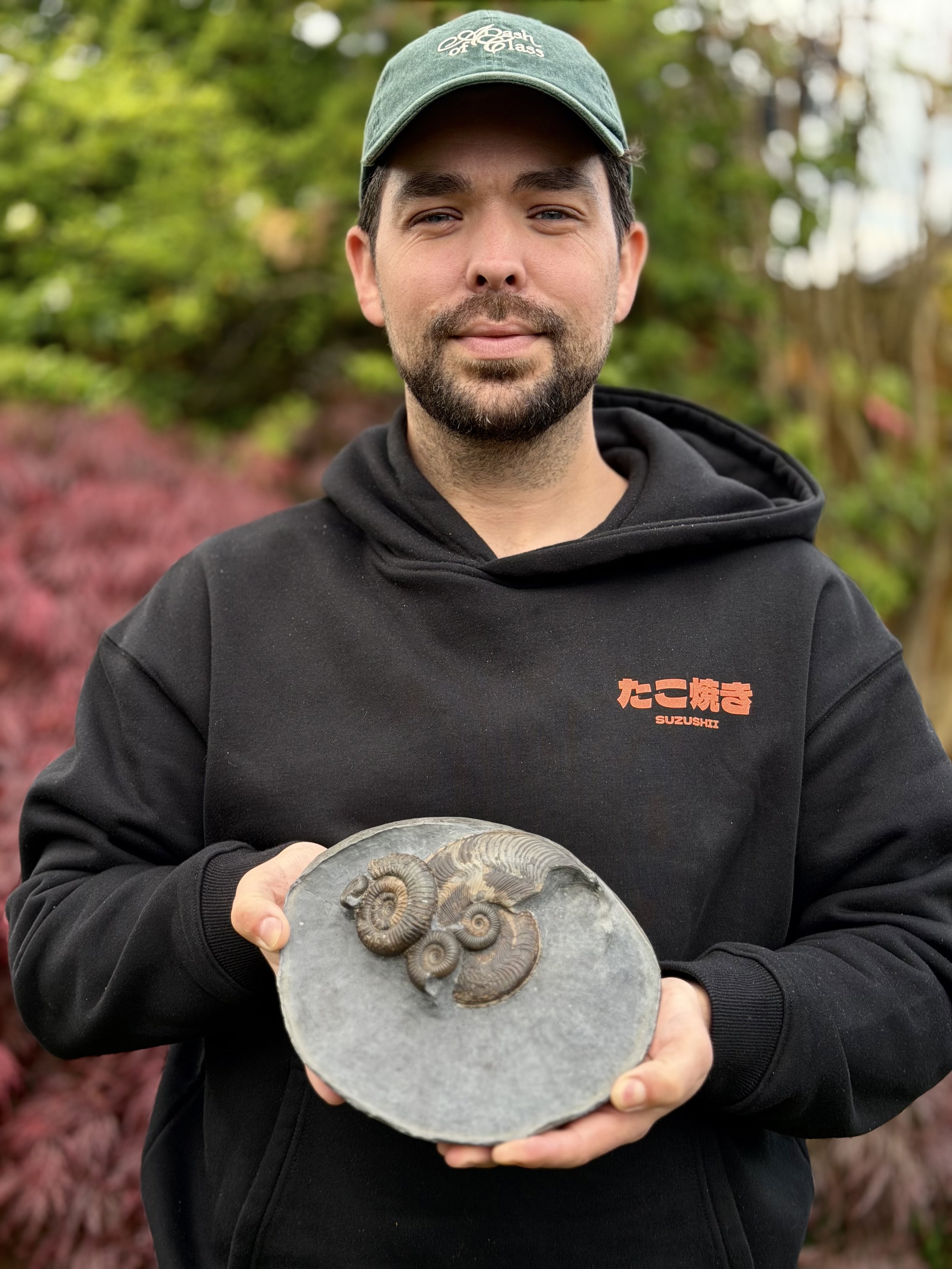 A man wearing a green cap and black hoodie holding a fossil of ammonite shells outdoors with blurred colorful trees in the background.