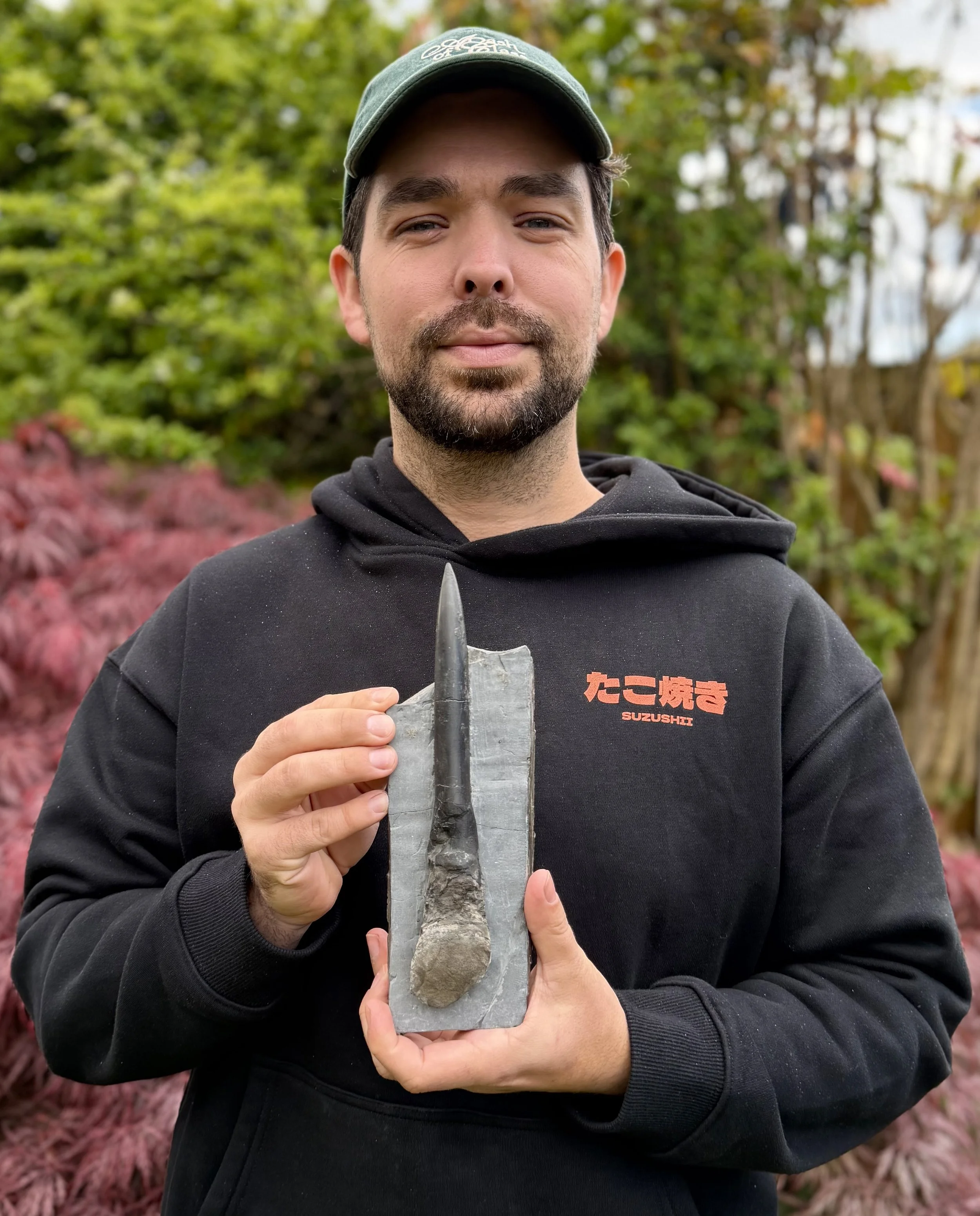 A man wearing a dark hoodie and cap holding a fossilized skull and jawbone of a prehistoric animal outdoors with green trees and bushes in the background.
