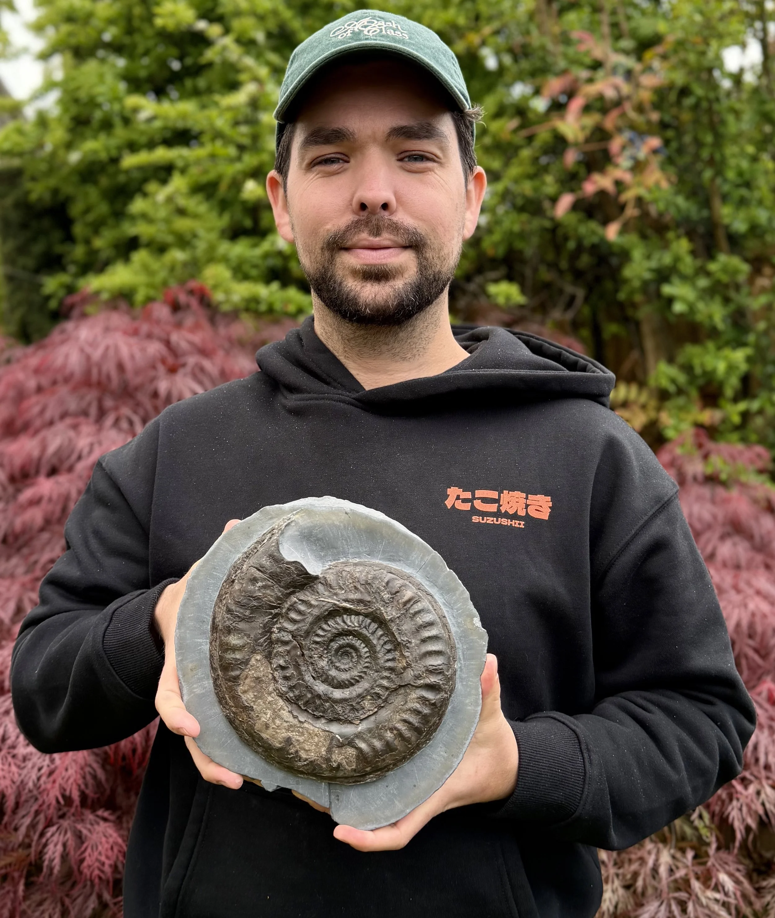 A man holding an ammonite fossil in front of colorful autumn trees, dressed in a black hoodie with orange Japanese characters and a green cap.