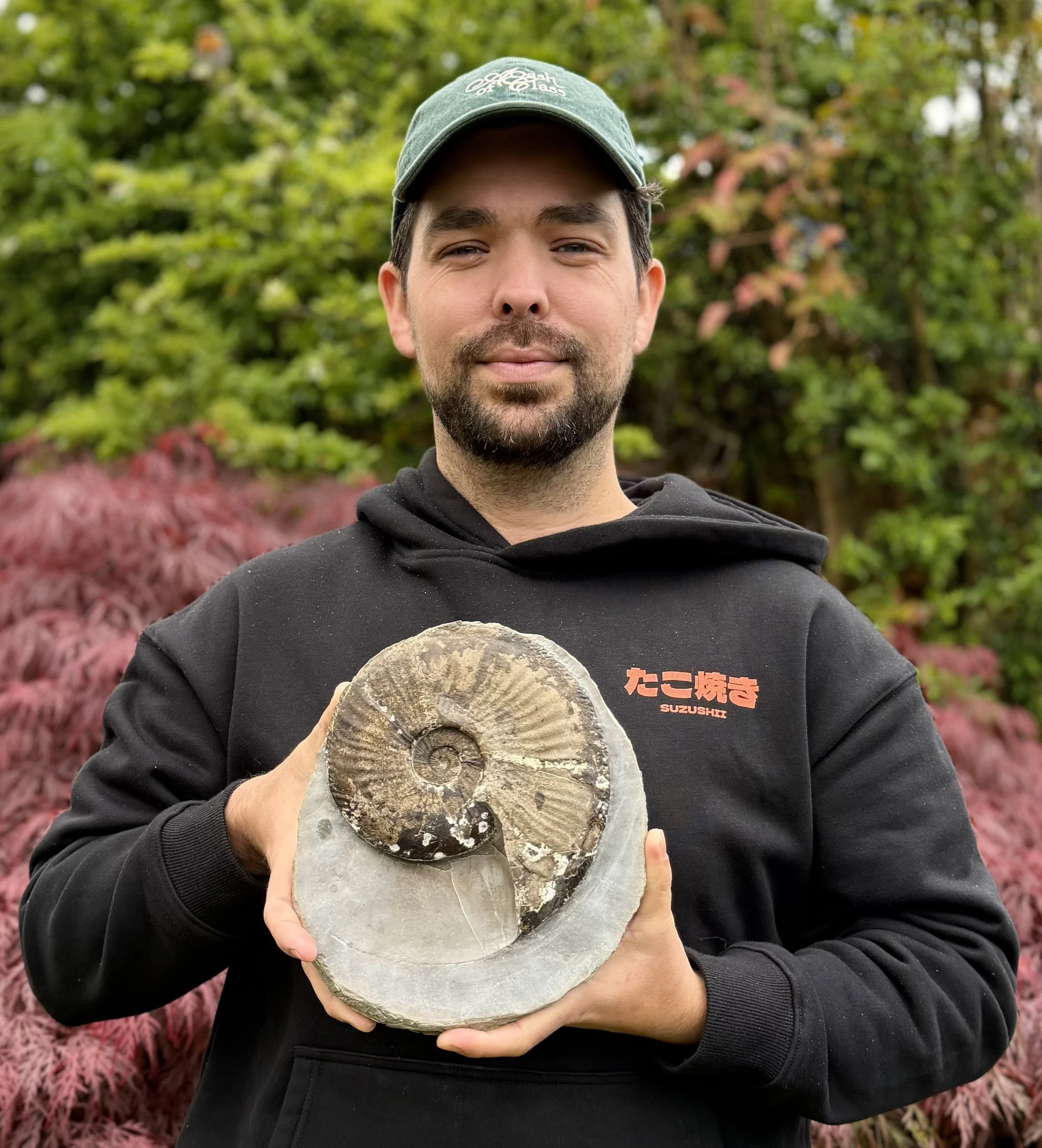 A man wearing a green cap and black hoodie holding a large fossilized ammonite shell in front of him outdoors, with a background of green and reddish foliage.