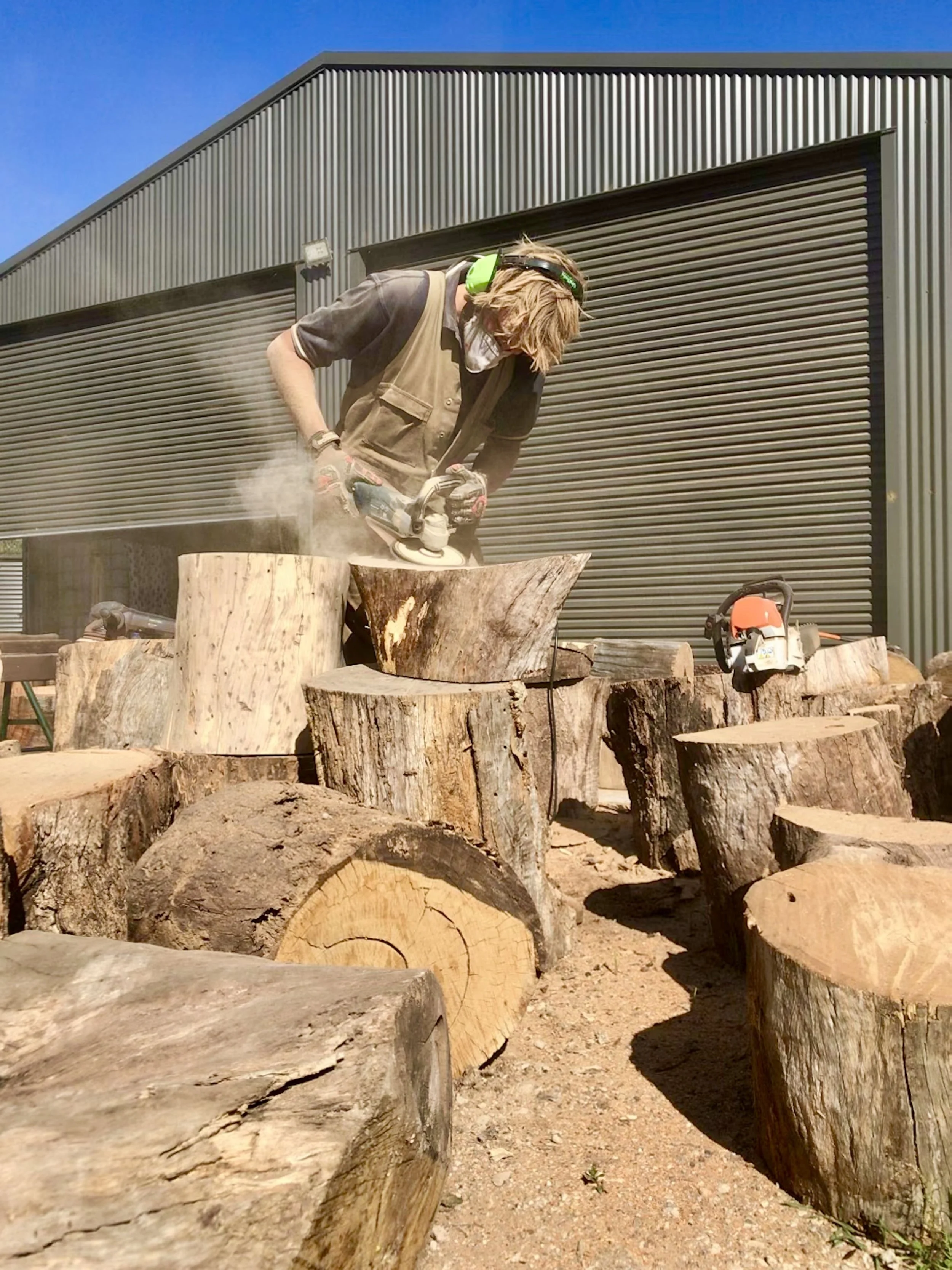Sam at Ugly Duckling Timber Co with safety earmuffs and gloves cutting a large piece of wood with a chainsaw outdoors, with scattered logs and shed in the background.