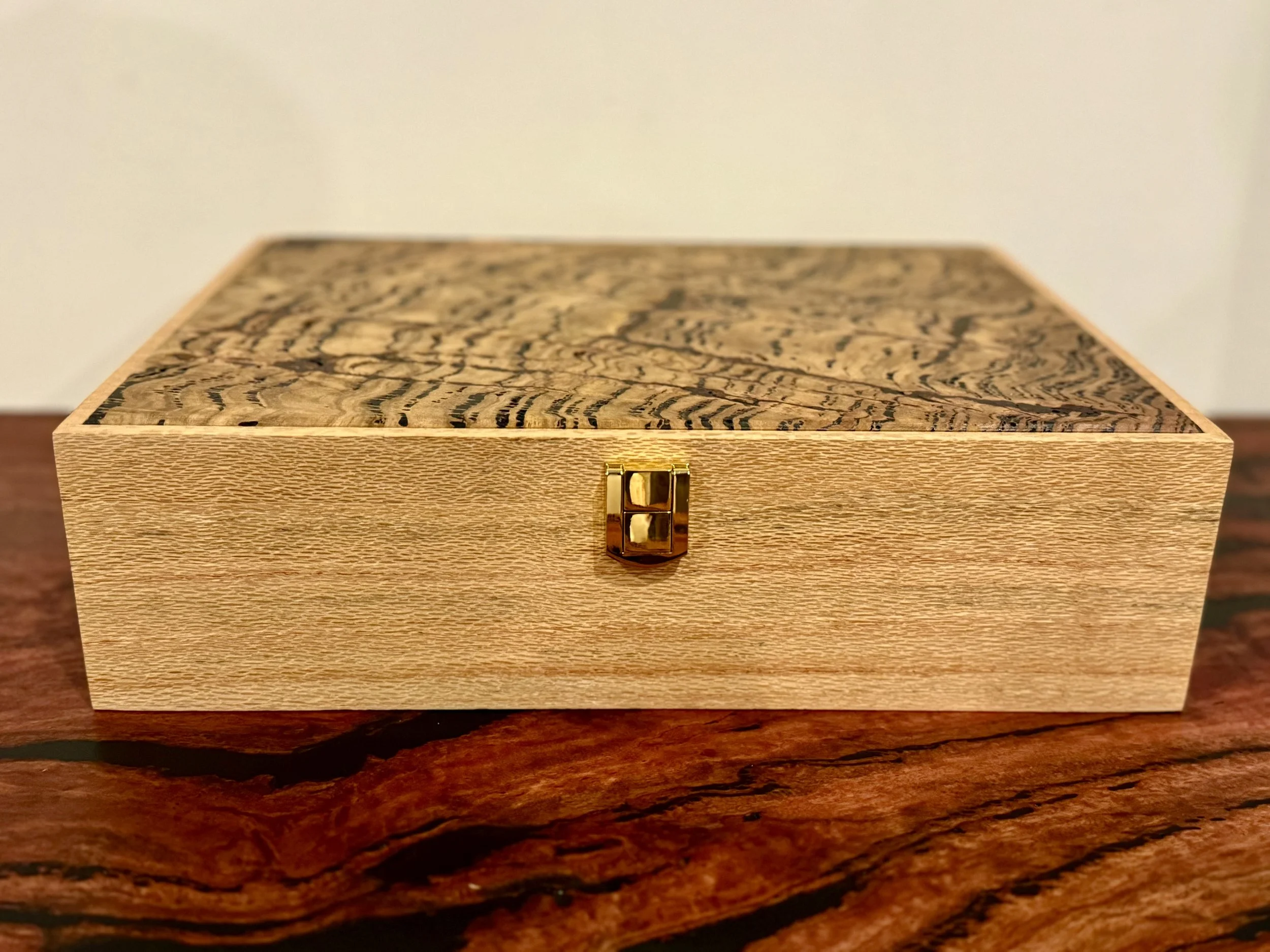 A rectangular wooden box with a decorative top and a small gold latch on the front, placed on a wooden surface.