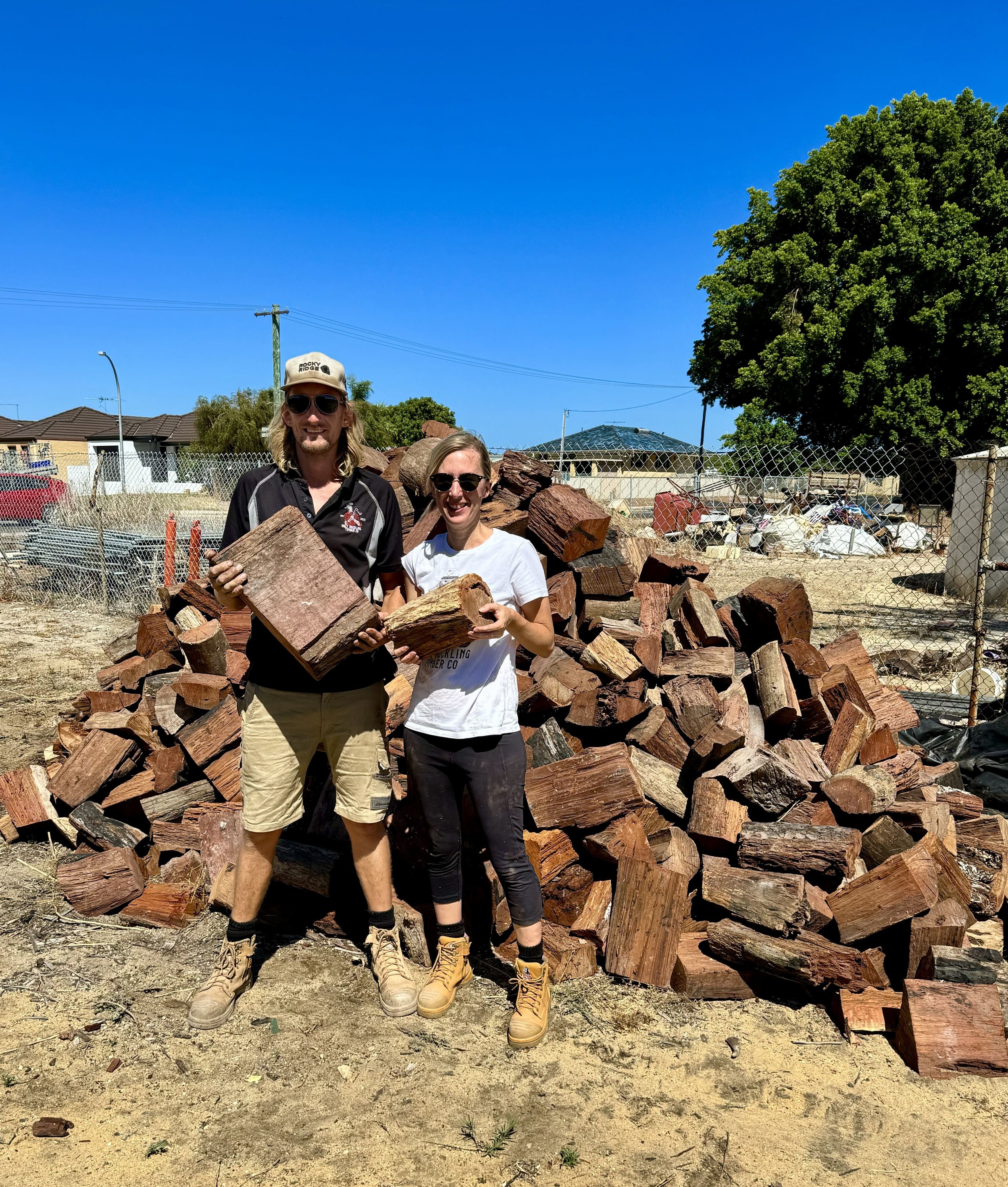 Sam and Katie, owners of Ugly Duckling Timber Co standing in front of a pile of cut firewood outdoors under a clear blue sky, holding pieces of wood and smiling.