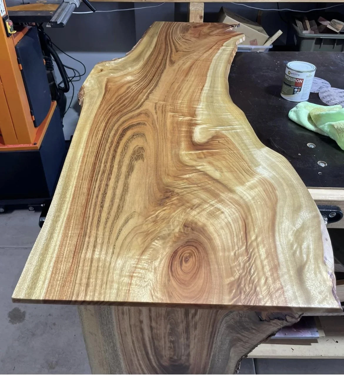 A large, polished slab of natural wood with rich grain patterns, placed on a workbench in a woodworking shop.
