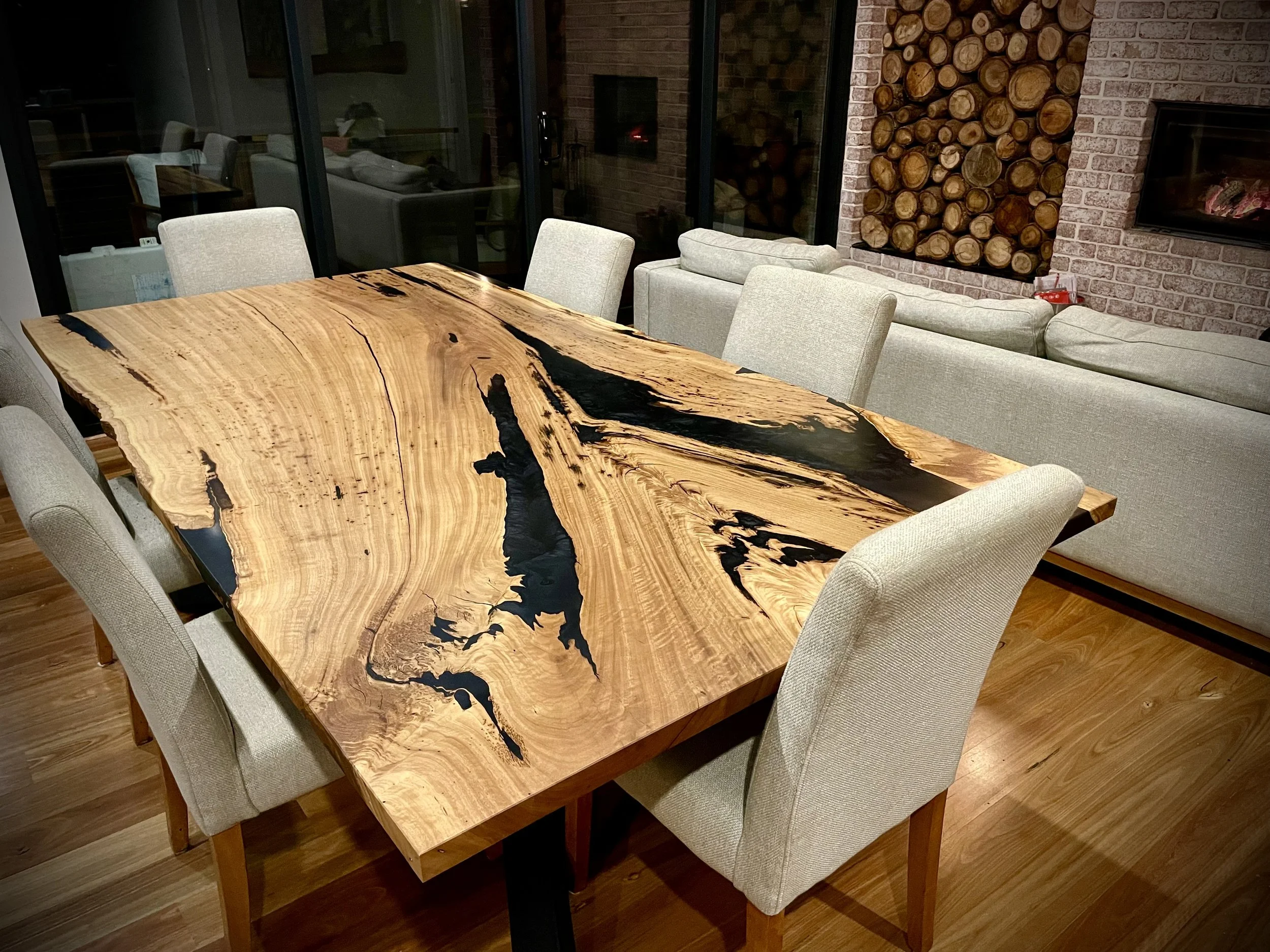 A wooden dining table with a natural edge and black resin accents, surrounded by beige upholstered chairs, in a modern dining room with brick walls and a stone fireplace