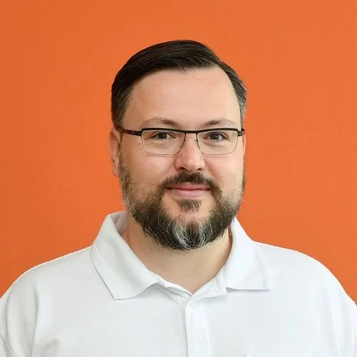 Ein Mann mit Brille, dunklem Haar, Bart und Schnurrbart trägt ein weißes Polo-Shirt vor einem orangefarbenen Hintergrund.