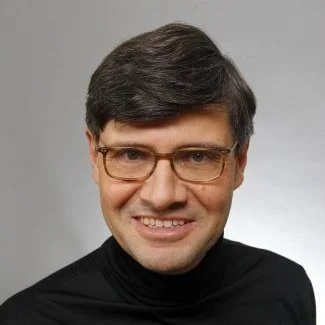 Ein Mann mit dunklem Haar, Brille und schwarzen Pullover lächelt vor einem grauen Hintergrund.
